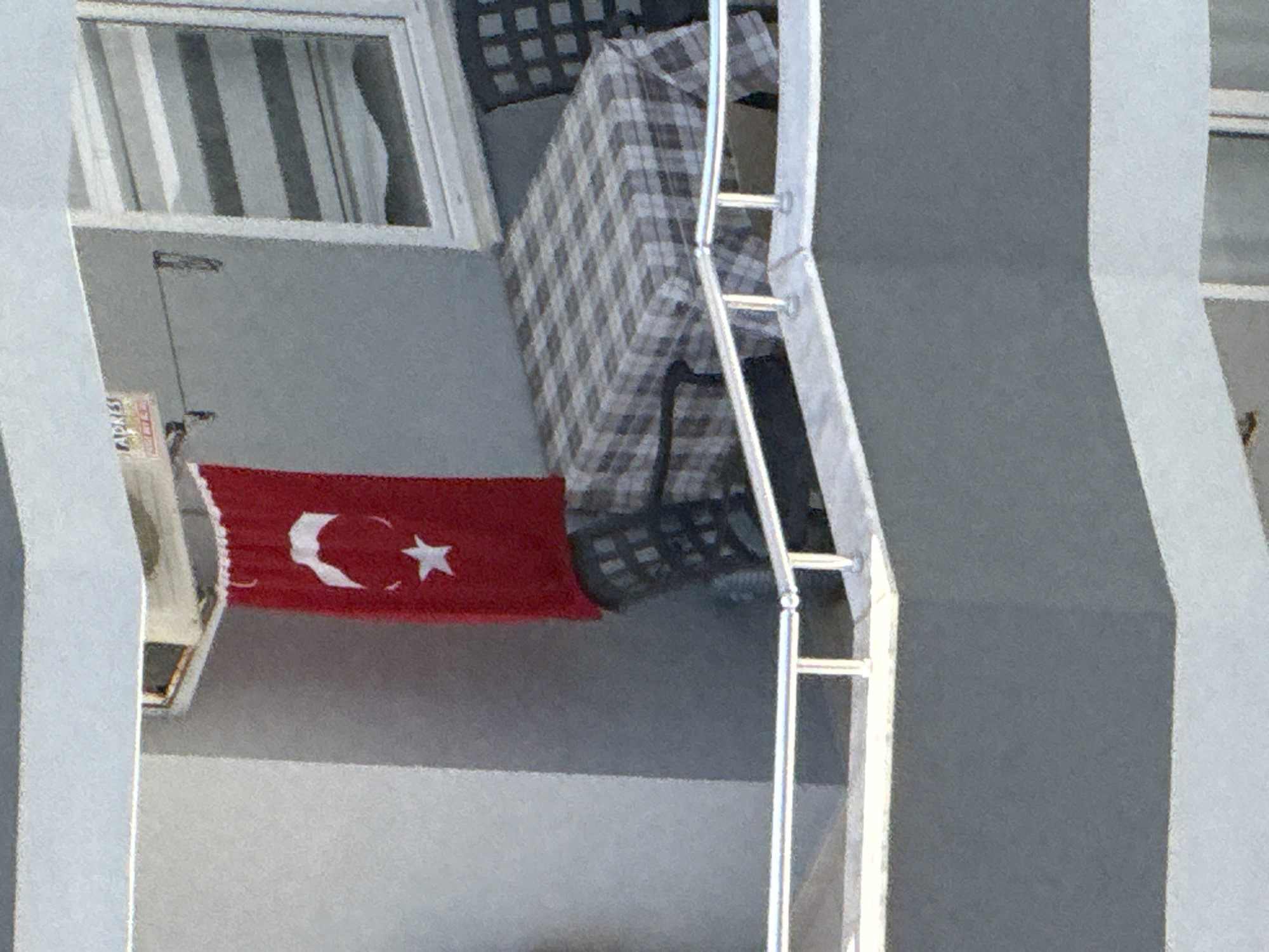 The image, when viewed in its correct orientation, depicts a segment of a building's exterior, focusing on a balcony and a window. A red flag, prominently featuring a white crescent moon and star, indicative of the Turkish national flag, hangs downwards from an attachment point on the balcony wall. To the left of the flag, what appears to be an outdoor air conditioning unit or similar appliance is visible. A thin cable or wire extends from near the window, downwards towards this unit.
On the balcony itself, a chair covered with a grey and white checkered (plaid) fabric is positioned. The balcony features a silver-colored metal railing. Above the balcony, a window is fitted with vertical blinds displaying a pattern of alternating light and dark grey stripes. The building's walls are painted in varying shades of grey and white.
The scene suggests a residential or urban setting during daylight hours, indicated by the bright, even lighting. No people or active events are visible. On a white label with a red stripe, located near the air conditioning unit, the word "ADRES" is faintly discernible.