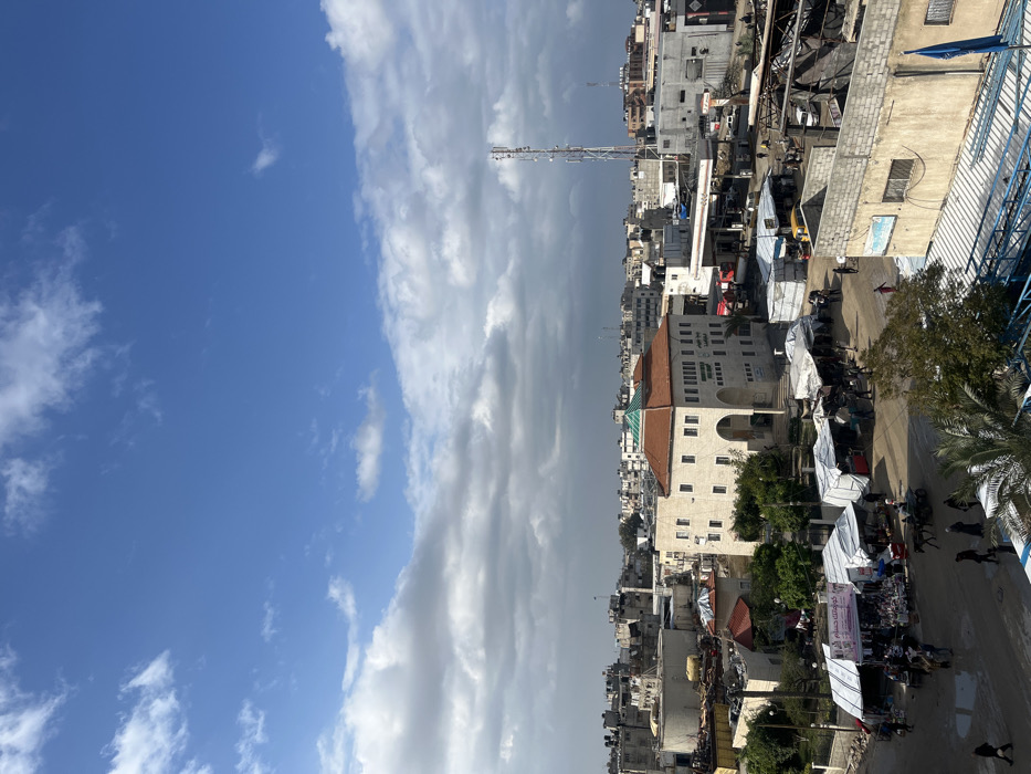 An elevated view captures an active urban scene in Khan Yunis, Palestinian Territory, under a partly cloudy sky with patches of blue. The central focus is a prominent light-colored, multi-story building with a distinctive reddish-brown tiled roof and an arched entrance, displaying Arabic text on its facade.
In the foreground and to the left of this central building, an open-air market operates, characterized by numerous stalls covered with white tarpaulins, with people congregated around them. A donkey-drawn cart is visible in this market area. The ground in the market appears unpaved, possibly muddy or damp.
To the right of the central building, an area resembling a gas station or vehicle service center is visible, featuring canopies and multiple vehicles, including what appear to be red trucks or emergency vehicles. Further right, a tall, white lattice communications tower rises above the surrounding buildings.
The broader urban landscape consists of densely packed, multi-story concrete and stucco buildings with flat roofs, extending into the distant hazy horizon. Visible within this dense urban fabric are areas of what appears to be rubble, damaged structures, and possibly charred debris, particularly in the mid-ground and left-hand side of the image. Large industrial-looking cylindrical tanks are situated in the upper-left background. Scattered individuals and small groups of people are visible traversing the streets and pathways. A blue flag is affixed to a pole on a building in the lower-right foreground. Sparse vegetation, including palm trees, is interspersed throughout the scene.
