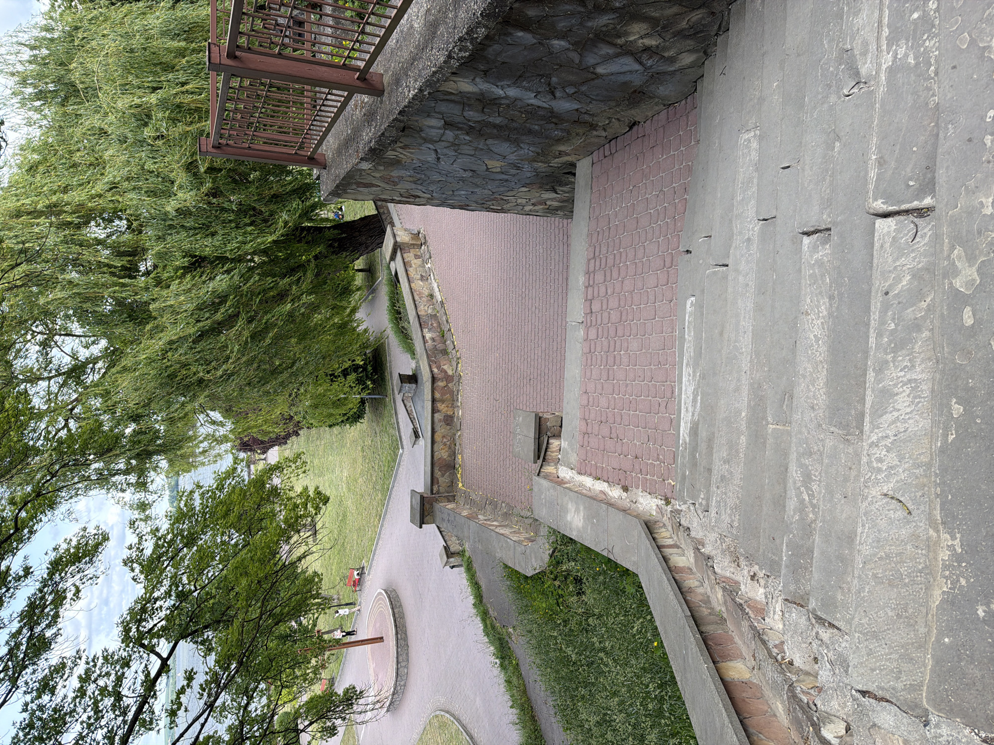 The image shows a park scene in Ternopil, Ukraine, photographed from an elevated position. A set of concrete stairs descends alongside a red brick path and a stone wall topped with a metal railing. Lush green trees frame the upper left of the image, partially obscuring the view of the park below. In the distance, a circular paved area with benches and people standing or sitting can be observed, along with grassy areas and a body of water in the far background. The photo seems to have been taken on a cloudy day with soft, diffused light.