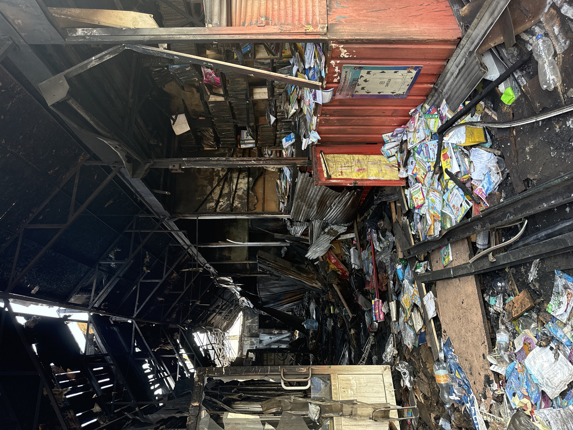 The image depicts the aftermath of a fire within what appears to be a bookshop or a similar retail establishment in Kyiv, Ukraine. The scene is one of extensive damage. The roof structure is severely compromised, with large sections missing, allowing sunlight to stream through gaps in the charred beams and sheeting. The interior is filled with debris, including scattered books, papers, and what appear to be plastic bottles and packaging. The walls and shelving units are blackened by smoke and fire damage. In the foreground, a large pile of books is visible, many of them appearing to be children's books with colorful covers, now mixed with ash and debris. To the right, a red corrugated metal structure with posters on it stands partially intact, contrasting with the surrounding devastation. The floor is covered in soot, ash, and scattered merchandise. The overall impression is one of a significant fire that has gutted the premises, leaving behind a chaotic and desolate scene.