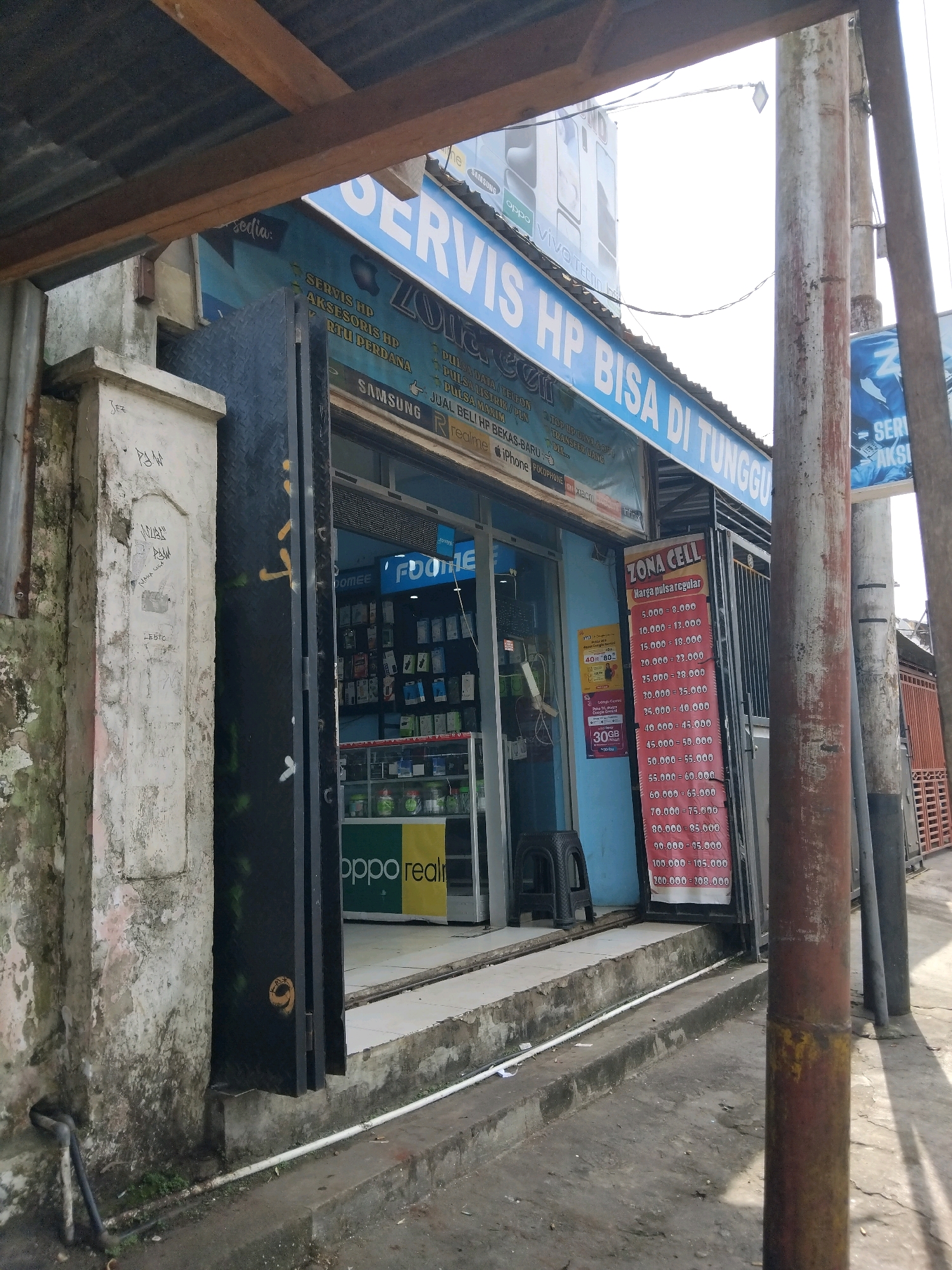 A storefront with a blue awning and a large sign reading "SERVIS HP BISA DI TUNGGU" is visible. The sign also features the text "ZONA CELL" and lists prices for phone services and data packages. Inside the store, shelves are stocked with mobile phones and accessories. A price list for phone credit is displayed vertically on the right side of the entrance. The exterior of the building shows signs of wear and tear, with peeling paint and graffiti on a pillar to the left. The surrounding area appears to be a street in a commercial district, with other buildings and utility poles visible in the background. The lighting suggests it is daytime.