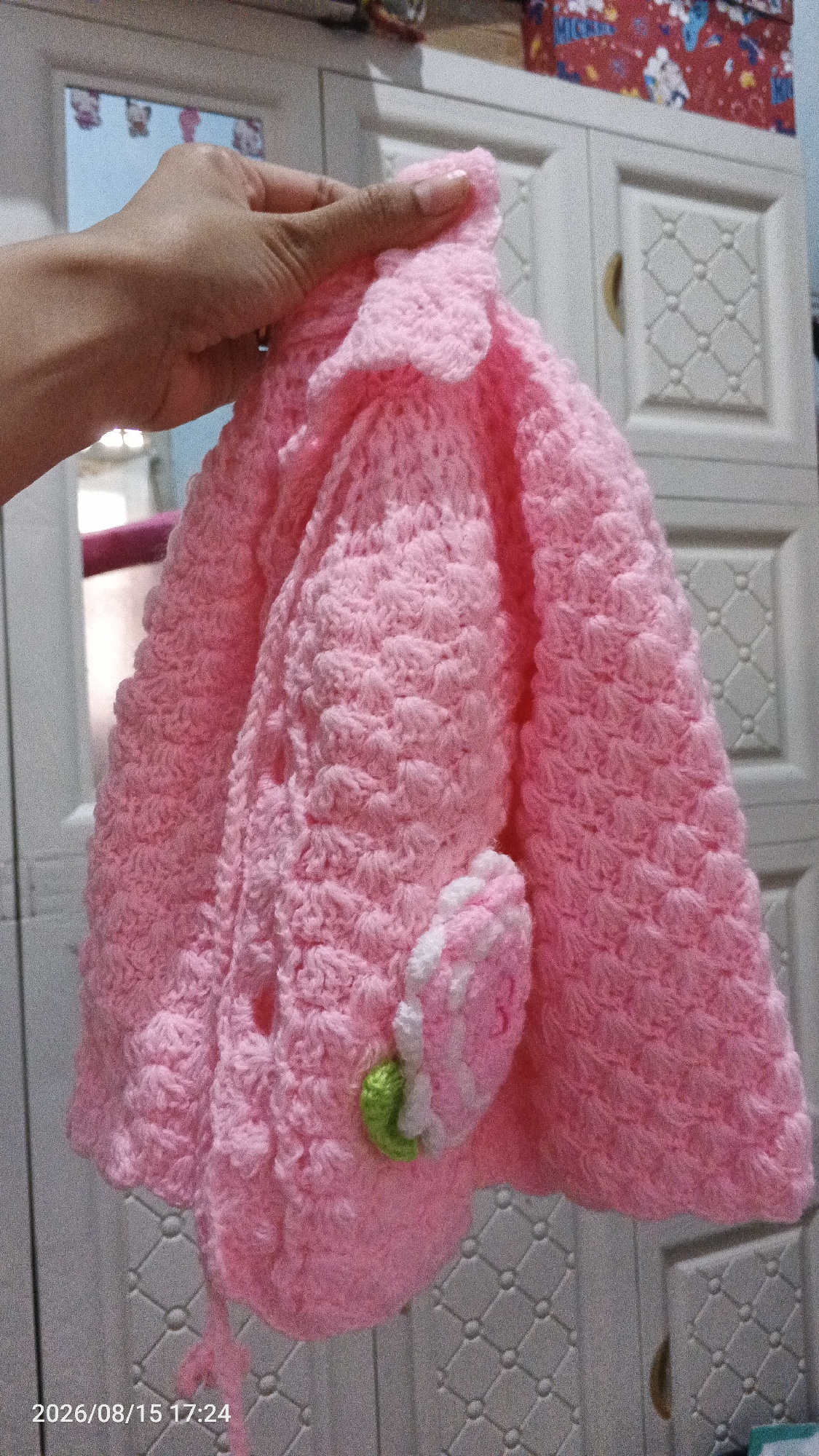 A hand holds a pink crochet item, possibly a small bag or purse, adorned with a crocheted pink and white flower with a green stem. The background features a white cabinet with a diamond pattern. The bottom left of the image shows a timestamp reading "2026/08/15 17:24". The context indicates this scene is located in Desa Rawajaya, Indonesia.
