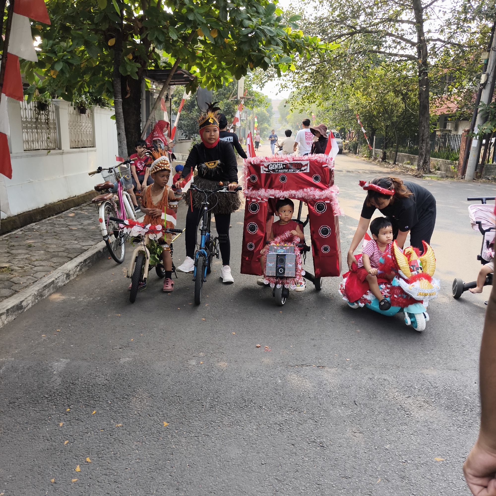 This image depicts a street scene in South Jakarta, Indonesia, likely during a local celebration or parade, given the presence of decorative elements and traditional attire. In the foreground, several children are participating. One child, wearing a traditional headdress and an orange vest adorned with fringes and armbands, sits on a small, decorated bicycle. Another child, dressed in a red outfit, sits in a red, tent-like structure mounted on a three-wheeled vehicle, which is decorated with what appear to be speaker motifs. A woman in a black shirt and a red headband is tending to this child. To the left, a child in a red shirt is seated on a bicycle. In the background, other people are visible, including individuals on bicycles and walking. Red and white Indonesian flags are displayed along the street, suggesting a patriotic or national event. The environment appears to be a residential area, with houses and trees lining the street. The asphalt road has some scattered dry leaves. The overall impression is one of community gathering and celebration.