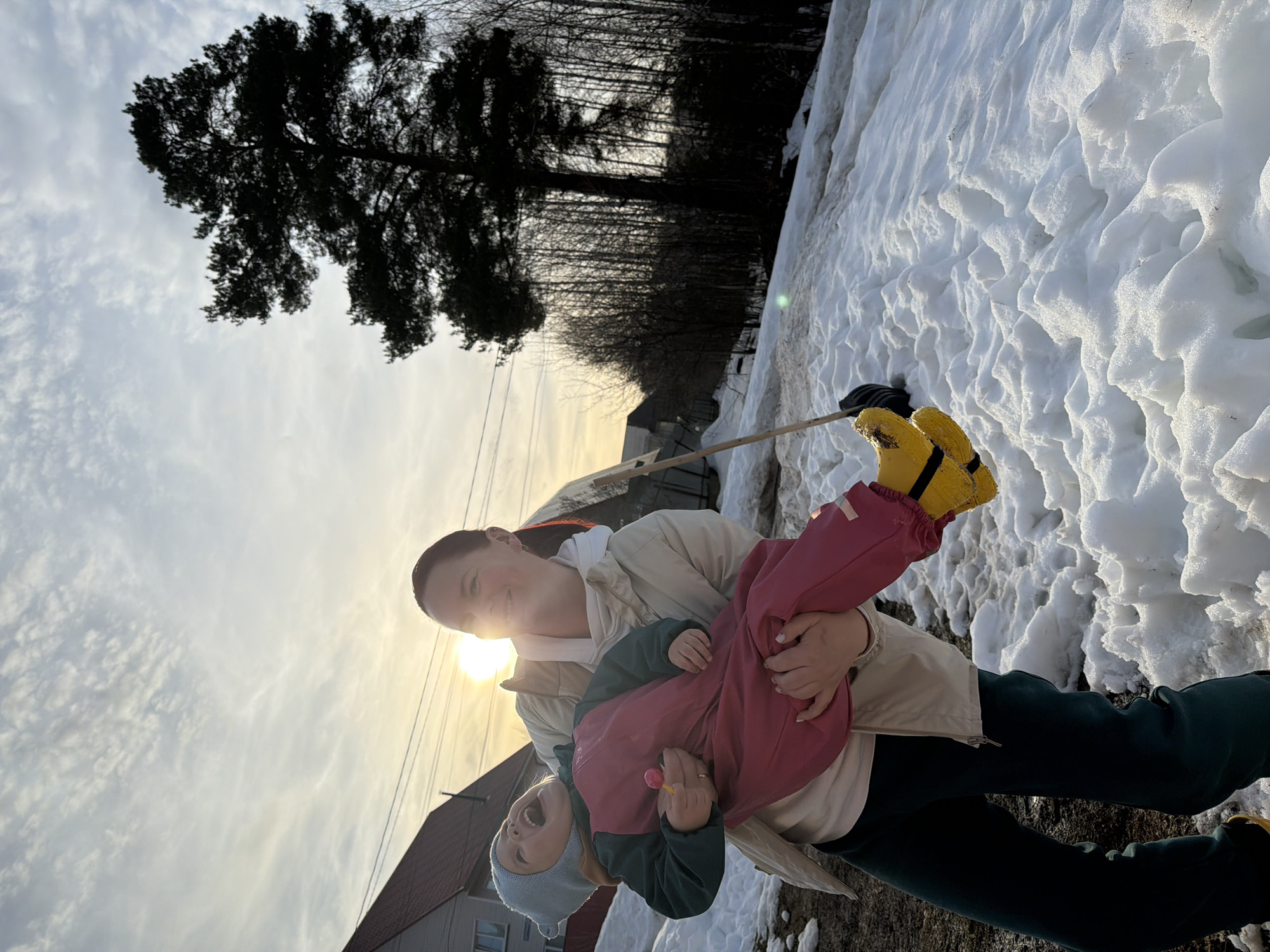 An adult female is depicted holding a young child horizontally in an outdoor, snowy environment in Chaikovsky, Russia. The adult, wearing a light-colored jacket, light top, and dark pants, has dark hair pulled back and is smiling towards the camera, partially obscured by sun glare. She wears a ring on her left ring finger. The child, dressed in a light blue beanie, dark green shirt, reddish-pink overalls, and bright yellow boots, is held across the adult's body, facing away. The child's mouth is open in an expression of joy or laughter, and a pink lollipop is held in their left hand.
The ground is covered in patchy, melting snow and exposed dark earth. In the background to the left, a two-story house with light siding and a reddish-brown gabled roof is visible, along with utility lines. To the right, a large, dark conifer tree stands prominently, backed by a dense stand of bare deciduous trees. A wooden stick with a dark implement (possibly a shovel head) is partially visible protruding from the snow near the right side. The sky is partly cloudy, and the scene is strongly backlit by a low-angle sun, suggesting late afternoon or early morning.