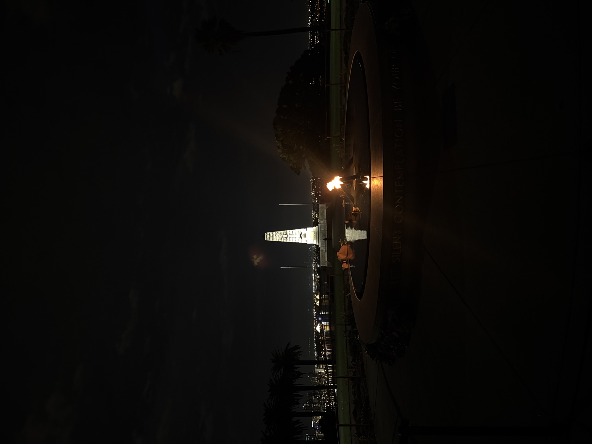 Nighttime view of the Flame of Remembrance at the State War Memorial in Kings Park, Perth, Australia. The flame is centered in a shallow, circular water feature with an inscription on the perimeter. In the background is a white stone cenotaph. Silhouetted palm trees are in the foreground. City lights stretch into the distance.