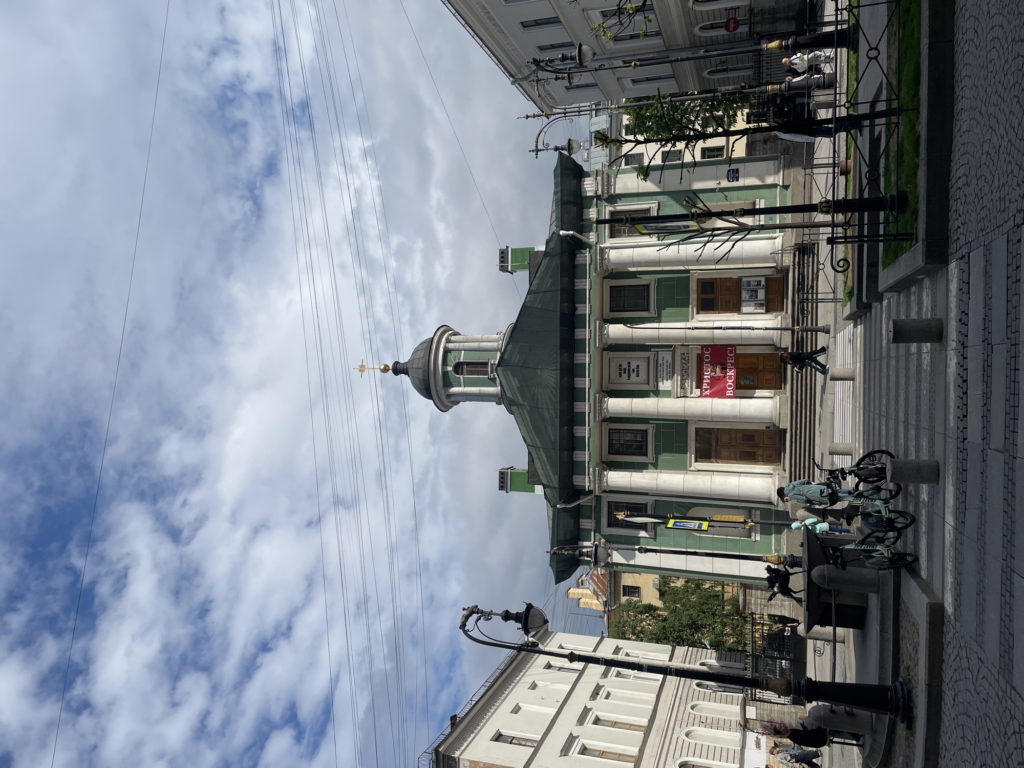 The image captures a view of the Church of the Savior on Sennaya Square in Saint Petersburg, Russia. The church has a green and white facade with a central dome topped by a cross. Its architectural style is characterized by columns.
Power lines stretch across the sky, which is filled with white clouds.
Several parked bicycles are located on the sidewalk.
People are present in the scene, including some on the steps leading up to the church.