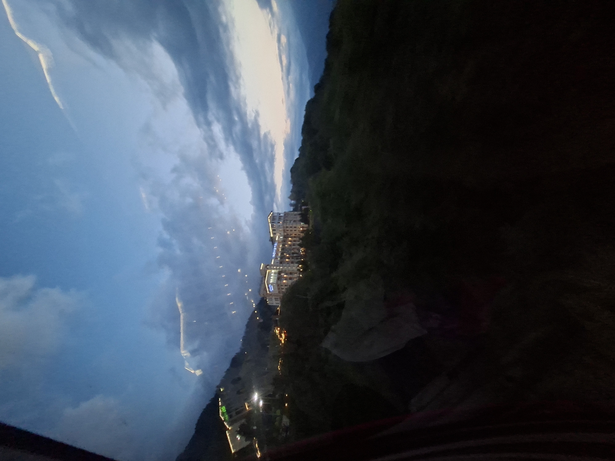 The media file is a photograph taken at dusk in Estosadok, Russia. The scene depicts a resort town nestled against a dark, forested mountainside. A multi-story hotel, illuminated by numerous lights, is prominently featured along the slope. The hotel's name, "NOVOTEL", is visible in neon blue lettering against its facade.
In the foreground, the image is partially obscured by what appears to be a dark, blurry object, possibly a blanket or a piece of clothing, and some streaks of color that could be from another fabric. The sky is a gradient of deep blue and twilight hues, with streaks of lighter clouds catching the fading light. Small, scattered lights from other buildings and streetlights dot the landscape, suggesting a bustling, illuminated town despite the approaching night.
The overall impression is one of a peaceful evening scene in a mountainous resort area, with the lights of the hotel and town creating a warm contrast against the darkening natural landscape and sky. The time of day is clearly twilight, as evidenced by the sky's color and the artificial lighting in the scene. The weather appears calm, with no strong indications of rain or wind. The location is identified as Estosadok, Russia, and the presence of the Novotel hotel serves as a specific landmark.