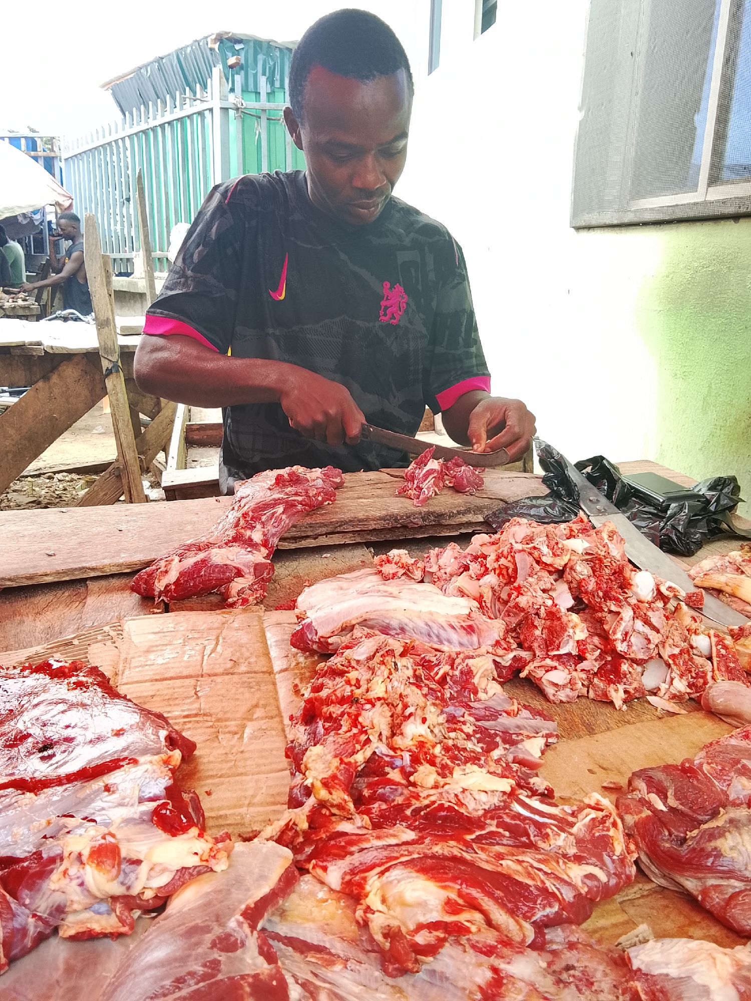 A butcher in Akwanga, Nigeria, is actively preparing cuts of raw meat on a wooden stall. He is a dark-skinned man wearing a black t-shirt with a pink trim and a pink Nike logo on the left chest. He is holding a knife in his right hand and using it to chop meat that is placed on a wooden board. In front of him, on the stall, are several large portions of raw beef, some already cut into smaller pieces, and a pile of chopped meat. A black plastic bag and another knife are also visible on the stall. The background shows a simple outdoor market setting with a corrugated metal fence and a light-colored wall. The overall scene depicts a typical day at a Nigerian butcher shop.