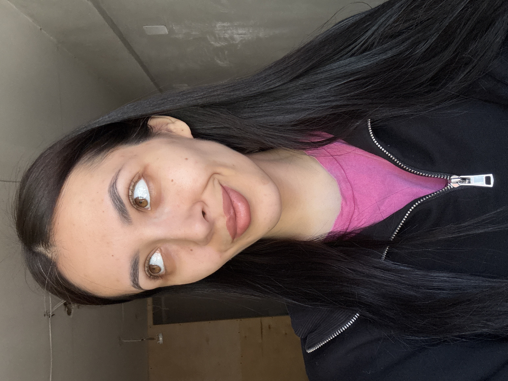 A woman with dark hair and brown eyes is pictured in a selfie-style image. She is wearing a pink top under a black zippered jacket. The background appears to be an unfinished interior space with grey walls and exposed construction elements. It's impossible to determine if the location is in Votkinsk, Russia, from the image alone.