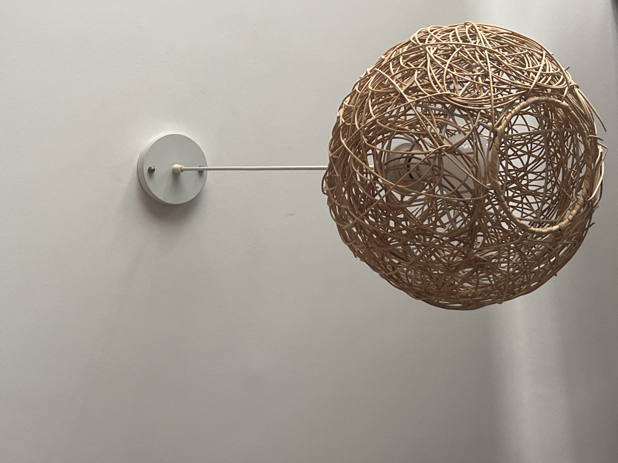 A spherical, woven lamp shade made of natural colored material hangs from a white ceiling. The lamp is suspended by a thin, white cord attached to a white circular fixture mounted on the ceiling. The woven material has an open, intricate pattern, allowing glimpses of the light bulb inside. The ceiling is a plain white surface with subtle shadows and variations in lighting, suggesting an indoor setting.