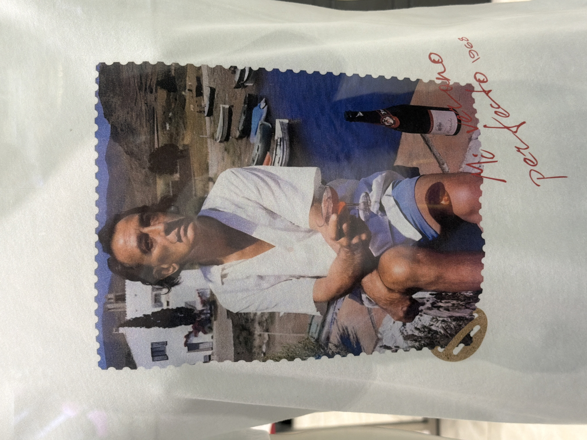 The image shows a postcard-style print of a man sitting by the water. He is fair-skinned with dark hair and is wearing a white shirt and blue swimming trunks. He is holding a glass in his left hand. Beside him is a bottle of what appears to be rosé wine. The background includes a body of water, boats, and buildings. Written in red ink on the white border of the print are the words "perfetto" and "Mio Veneto 1968". A perforated edge surrounds the print, giving it the appearance of a stamp or postcard. The image is presented on a white surface. There is no indication this is set in Novoaleksandrovka, Russia.