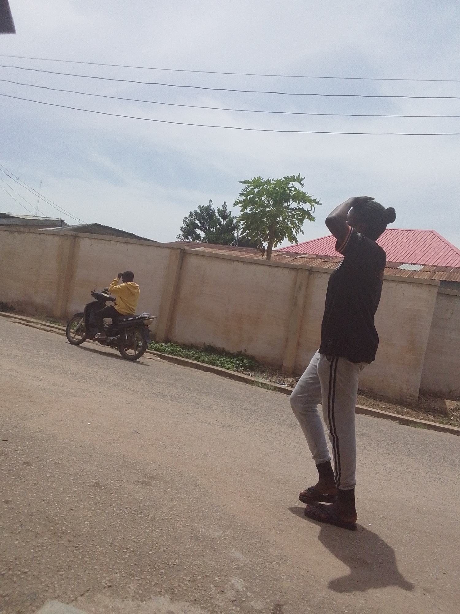 This image captures an outdoor street scene on a bright, sunny day. In the foreground, on the right, a person stands on an asphalt road with their back partially to the viewer. They are wearing a black short-sleeved top, light grey track pants with dark stripes down the sides, and dark open-toed sandals over black socks. Their head is tilted back, and their hands are raised towards their head, possibly adjusting hair or shielding their eyes from the sun. A distinct shadow is cast by this person onto the road.
In the midground, to the left, another individual is seated on a dark-colored motorcycle. This person is wearing a bright yellow hooded top and dark trousers. They also appear to be looking upwards or away, with their hands near their head. The motorcycle is stationary on the paved road, which seems to have a gentle incline.
Behind the subjects, a long, light-colored wall extends across the frame. Above the wall, green trees and foliage are visible, along with parts of buildings, including one with a prominent red corrugated metal roof on the right. Overhead, multiple parallel power lines stretch across a clear, light blue sky with some wispy clouds. The strong shadows on the ground suggest it is mid-day. No visible text is present in the image.
