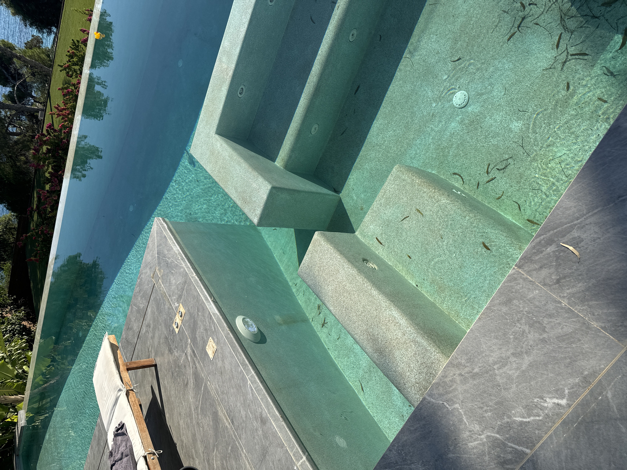 The image shows a view of a swimming pool, likely in Vouliagmeni Municipal Unit, Greece, given the context of the surrounding landscape and water. The pool features an infinity edge, with clear turquoise water. Built-in steps with a textured finish lead down into the pool. The poolside is paved with large, dark gray stone tiles. On one side, there is a built-in seating area or shallow section of the pool. A wooden lounger with a white cushion and a dark gray towel is placed on the tiled deck. The water contains some debris, appearing to be leaves or plant matter. In the background, lush green foliage and trees are visible, with a glimpse of the sea and a bright blue sky. Sunlight casts shadows across the pool area.