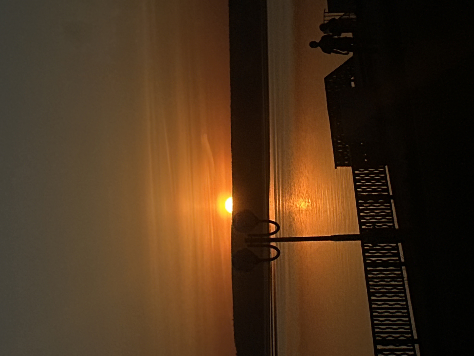A vertical shot captures a sunset over water. The sun, a brilliant orange orb, sits on the horizon, its reflection shimmering on the water's surface. To the right, a dark structure stands silhouetted, likely a pier or boardwalk, with a discernible railing pattern. Two figures are visible on the structure, their silhouettes suggesting they are observing the sunset. A lamppost stands near the water's edge, its curves echoing the setting sun. The sky is a gradient of orange, fading into darkness. The location is likely Votkinsk, Russia, given the information provided.