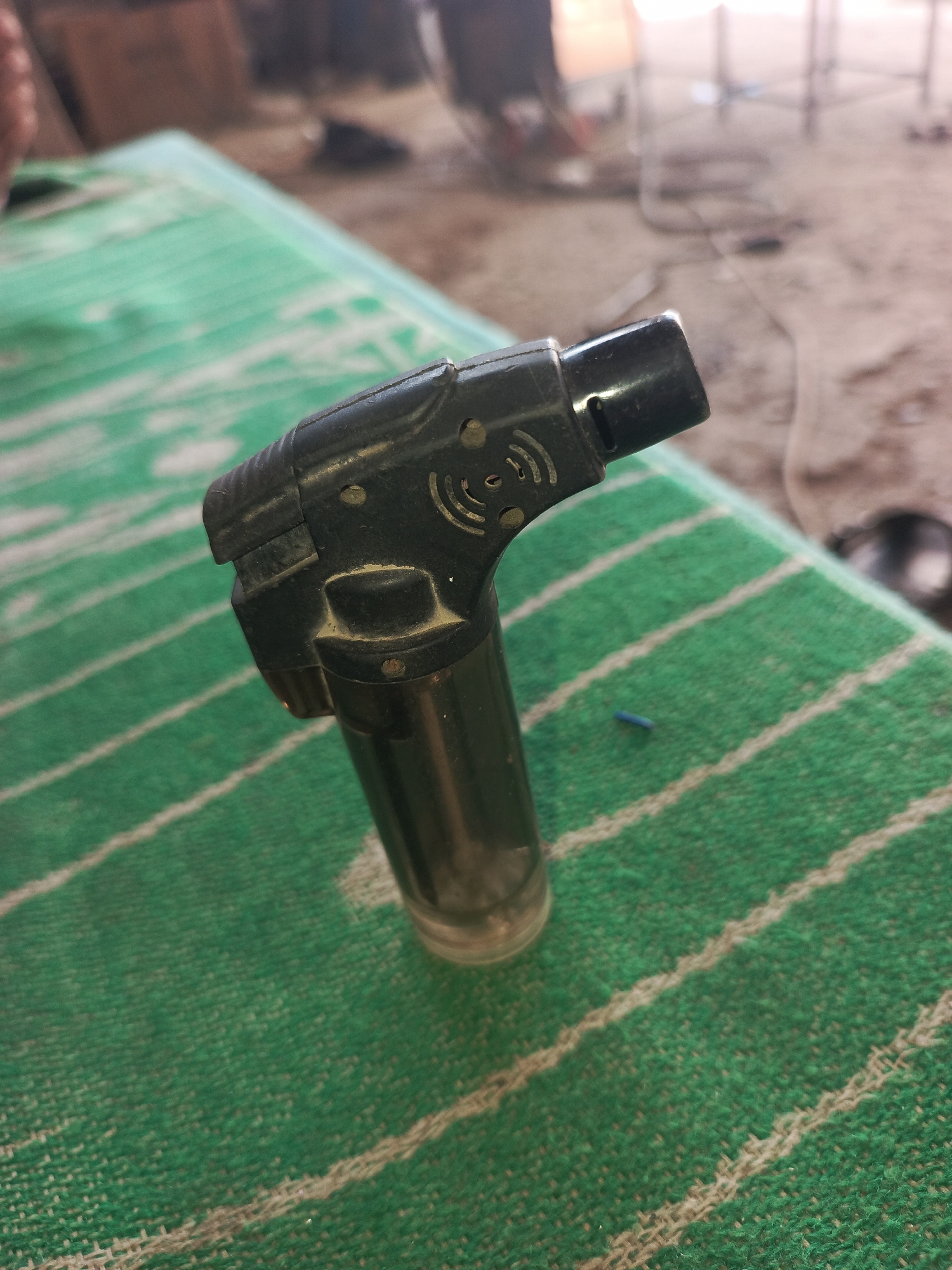 A black butane torch is positioned diagonally on a green and white striped cloth. The torch has a metallic sheen and appears to be made of plastic and metal. The background is blurred, showing what appears to be an outdoor or workshop setting with dusty ground and some indistinct structures. There are no people visible in the immediate vicinity of the torch. The image is taken from a slightly elevated angle, looking down at the torch. The location is specified as Kumkurgan, Uzbekistan.