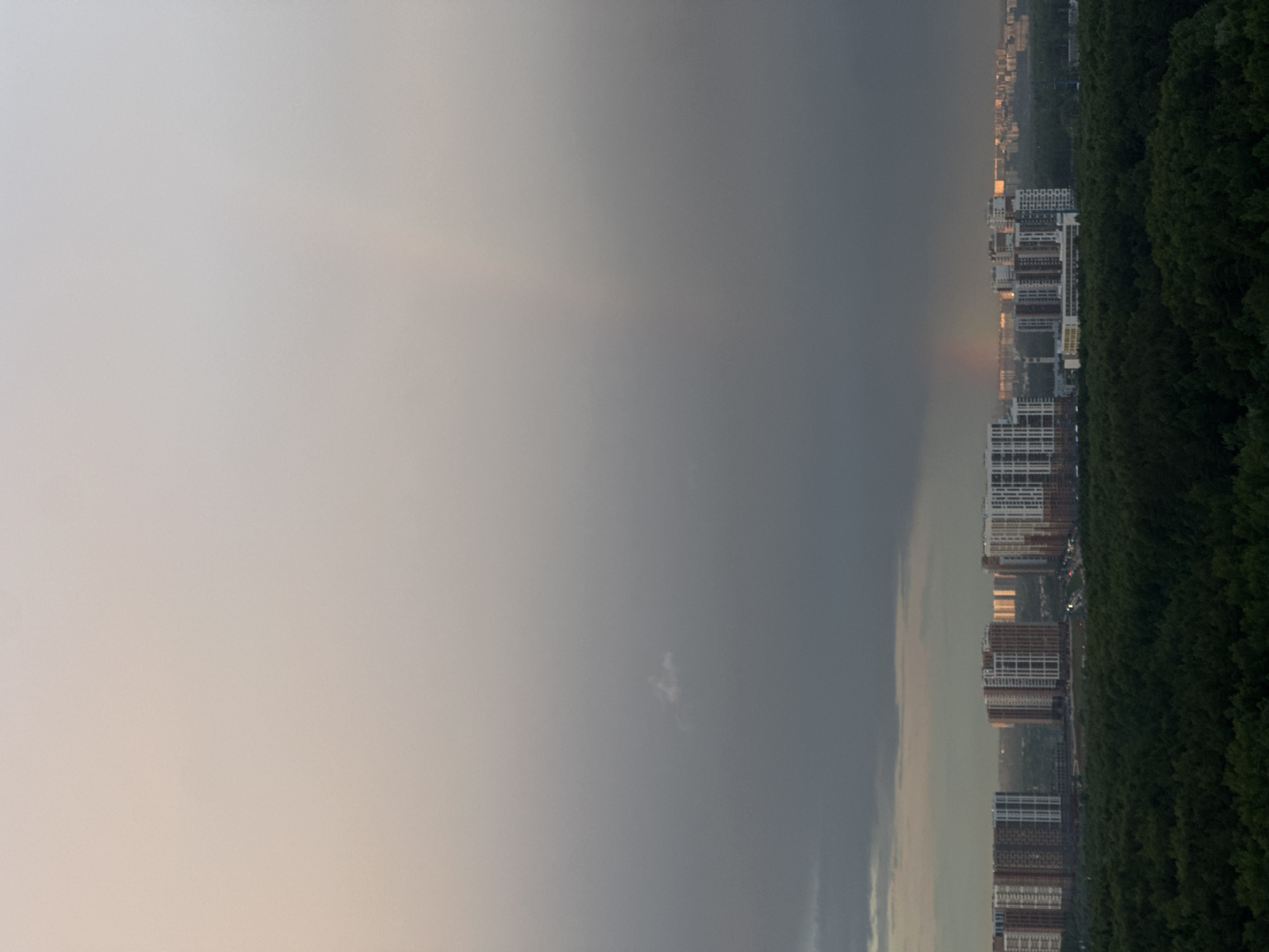The image shows a view over a city with tall residential buildings along a tree-lined edge. To the left of the buildings, the sky transitions from a brighter, overcast area to a darker, stormy-looking section. The city's location is Unknown, Unknown.