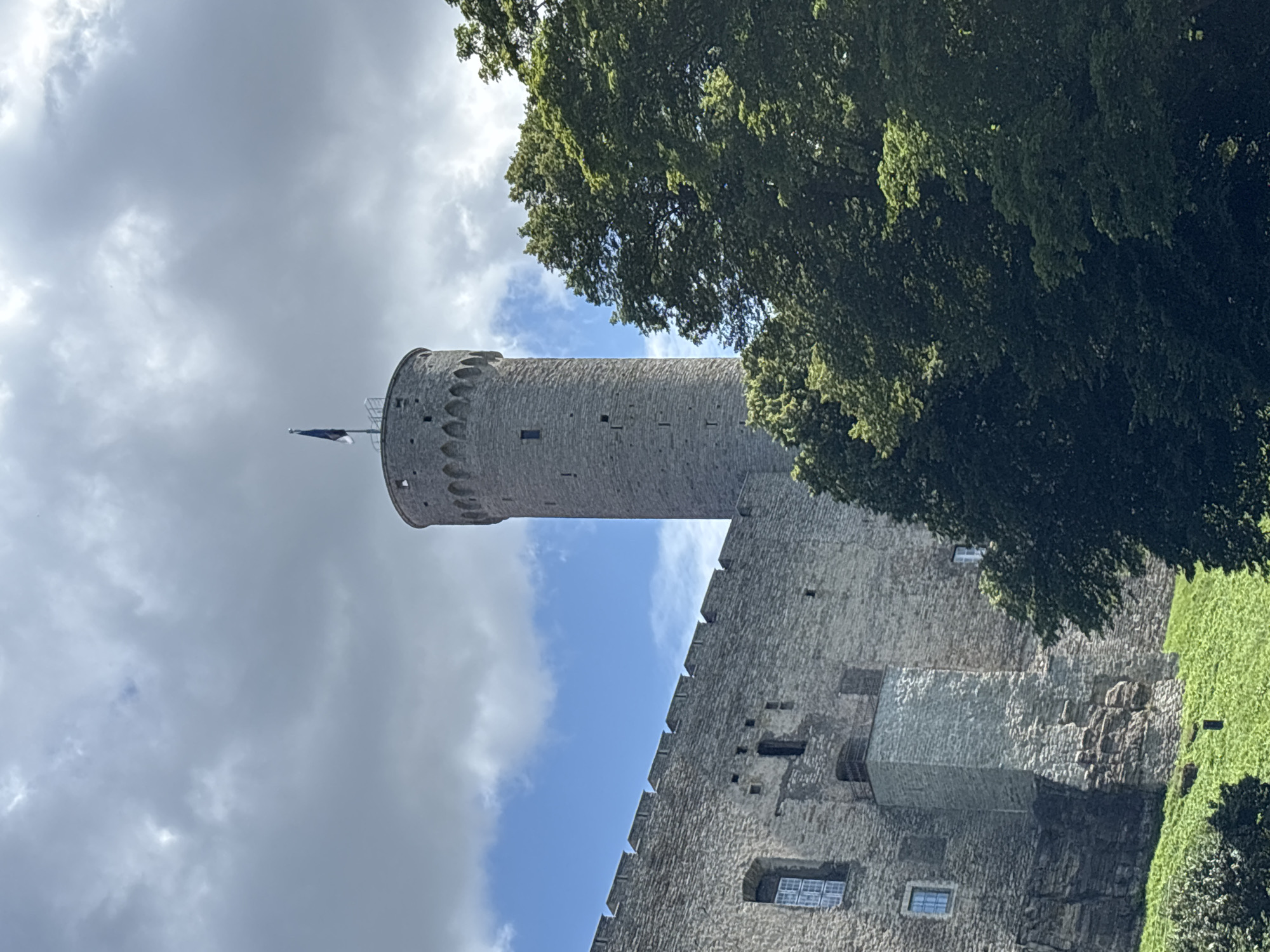 The image shows the Tallinn City Tower, also known as Pikk Hermann, a prominent defensive tower of Toompea Castle in Tallinn, Estonia. The tower is a tall, cylindrical stone structure with a flag flying at its peak. The castle walls, made of similar stone, extend to the left of the tower. The sky is partly cloudy with patches of blue sky visible. Lush green trees are visible in the foreground and to the right of the tower, obscuring parts of the castle. The sun is shining, casting light and shadows on the stone structures and trees. The overall scene depicts a historic landmark on a bright, partly cloudy day.