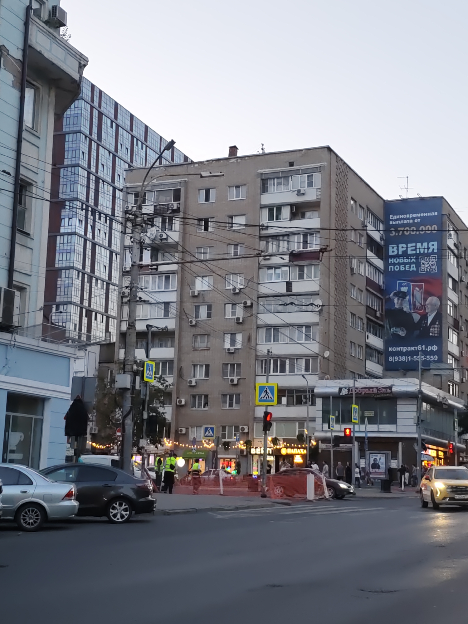 The image captures a street scene in Rostov-on-Don, Russia. In the foreground, several cars are parked or in motion on a paved road. A pedestrian crossing is visible, with traffic lights indicating a red signal. On the right side of the road, a light-colored car is turning.
Further back, a modern glass-fronted building stands tall, contrasting with older, multi-story residential buildings. One of these buildings features a large blue advertisement banner displaying Russian text and imagery of two men in uniform. The banner has the words "ВРЕМЯ НОВЫХ ПОБЕД" (Time for New Victories) prominently displayed.
On the sidewalks and near the buildings, there are people dressed in everyday attire, some appearing to be pedestrians and others possibly workers, given the high-visibility vests worn by a few individuals. A utility pole with numerous wires is a noticeable feature in the mid-ground, connecting various parts of the urban infrastructure. The overall environment suggests a typical city street with commercial and residential elements.