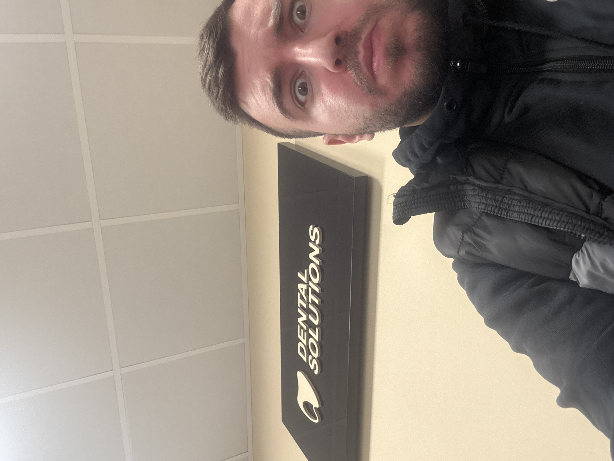 A selfie of a male with short brown hair and a beard, possibly in his late 20s or early 30s, wearing a black jacket. He appears to be inside an office building or clinic in Sofia, Bulgaria. Behind him is a sign that reads "DENTAL SOLUTIONS" with a logo resembling a tooth. The wall is beige and there is a suspended grid ceiling in the background.