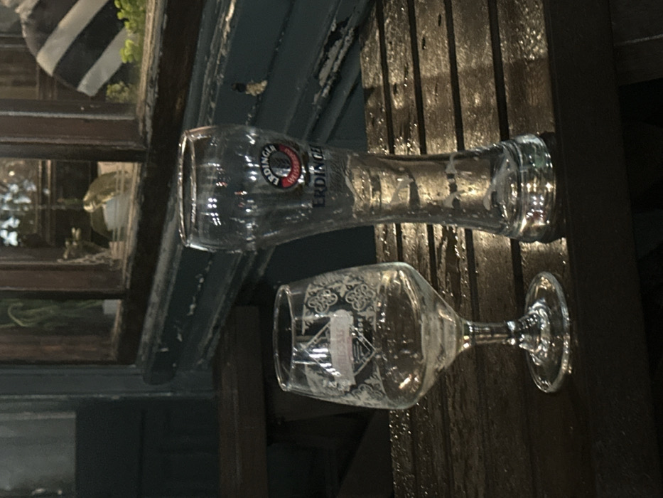 Two empty beer glasses sit on a dark wooden surface. The top glass is a tall, slender pilsner glass with the "Erdinger Weissbier" logo. Below, a smaller wine glass with a stemmed base displays ornate designs on the bowl. The glasses are positioned at an angle, partially obscuring a window frame and a striped cushion in the background. The scene is dimly lit.