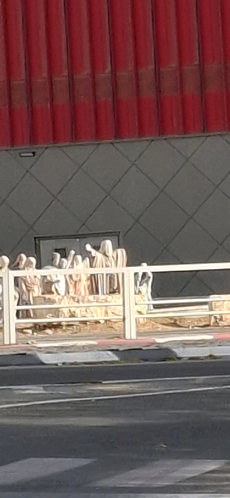 The image captures a scene in Be'er Sheva, Israel, with a prominent red corrugated facade visible at the top. Below this, a dark gray wall with a diamond pattern forms the background. In the middle ground, a group of light-colored statues, appearing to be people in robes, are arranged behind a white railing. One statue on the far right has a black and white pattern on its lower half, resembling a dog. A closed doorway with two panels is situated behind the statues. The foreground shows a street with asphalt and a white painted curb. The overall impression is of an outdoor public space, possibly a walkway or plaza.