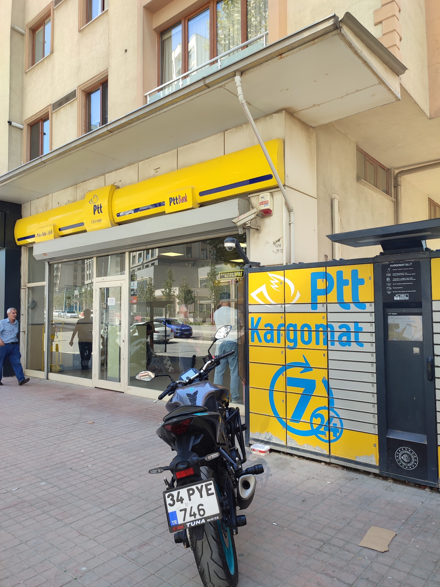 The image depicts the exterior of a PTT (Turkish Post) bank and PttKargomat (parcel locker) location on a sunny day in Kadıköy, Turkey. The building features a prominent yellow awning with the Ptt and PttBank logos. Below the awning is a glass-fronted entrance to the PTT bank. To the right of the bank entrance is a large yellow PttKargomat unit with multiple lockers and the Ptt logo and "Kargomat" text. A blue "7/24" logo is visible on the Kargomat.
In the foreground, a black motorcycle with turquoise accents is parked on a red brick sidewalk. The motorcycle has a license plate reading "34 PYE 746 TR TUNA". A man in a blue plaid shirt and dark pants is walking away from the camera on the left side of the sidewalk. Inside the PTT bank, several people are visible, including one person near the entrance and others further inside. Reflections in the PTT bank windows show trees and buildings.
The environment appears to be an urban street with buildings lining the sidewalk. The overall impression is that of a functional, active public service location.