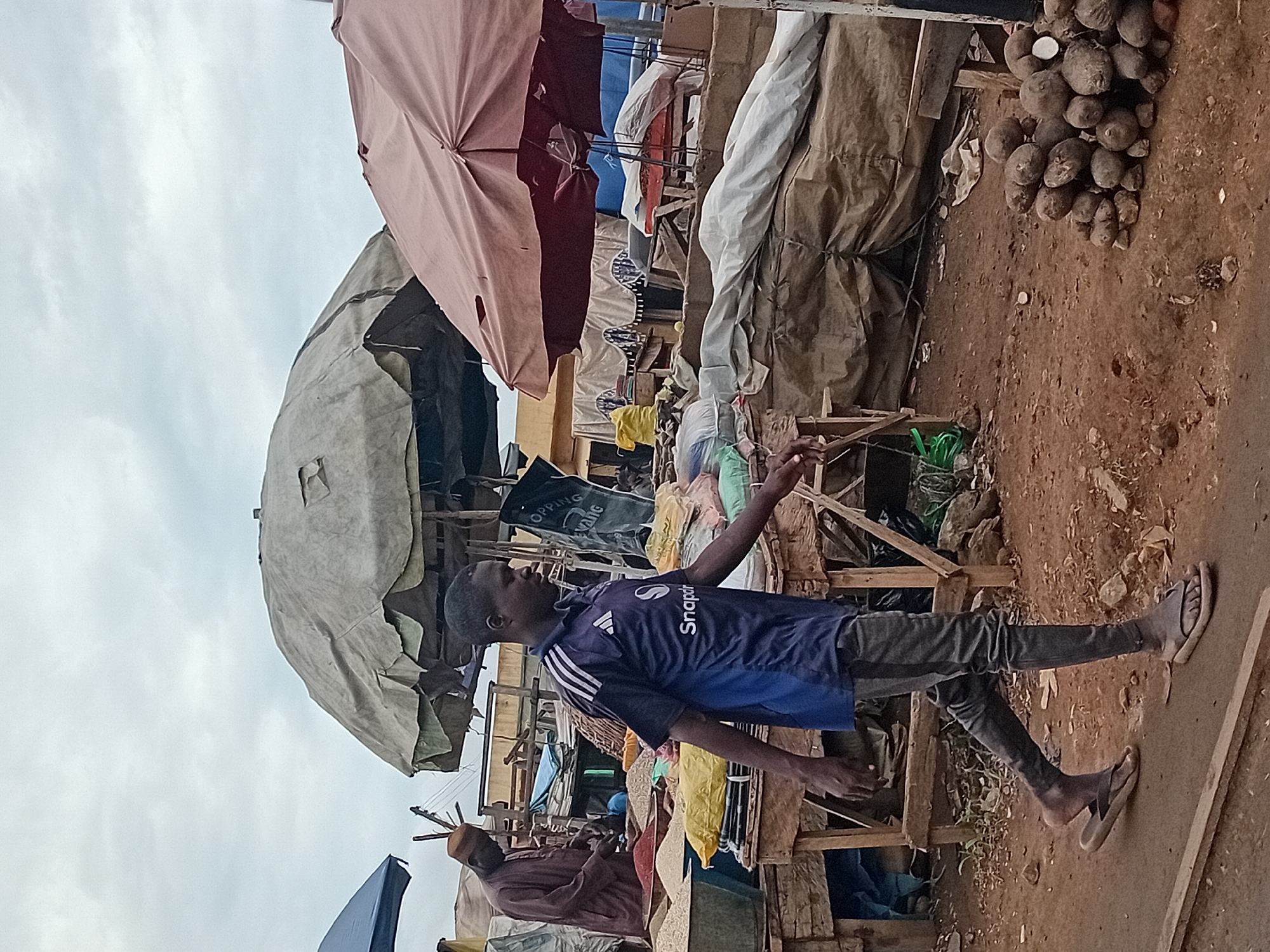 This image depicts an outdoor market scene in Maiburiji, Nigeria. A young man wearing a blue shirt with white stripes and dark pants, along with dark sandals, is walking in the foreground. He is looking towards the right side of the frame. Behind him, a man in a maroon robe and cap is tending to goods on a table. The market is characterized by various stalls covered by large, worn umbrellas. Some stalls display sacks and piles of what appear to be grains or spices. In the right foreground, a pile of yams is visible on the dusty ground. The environment is earthy with some vegetation and debris scattered around. The sky is overcast.