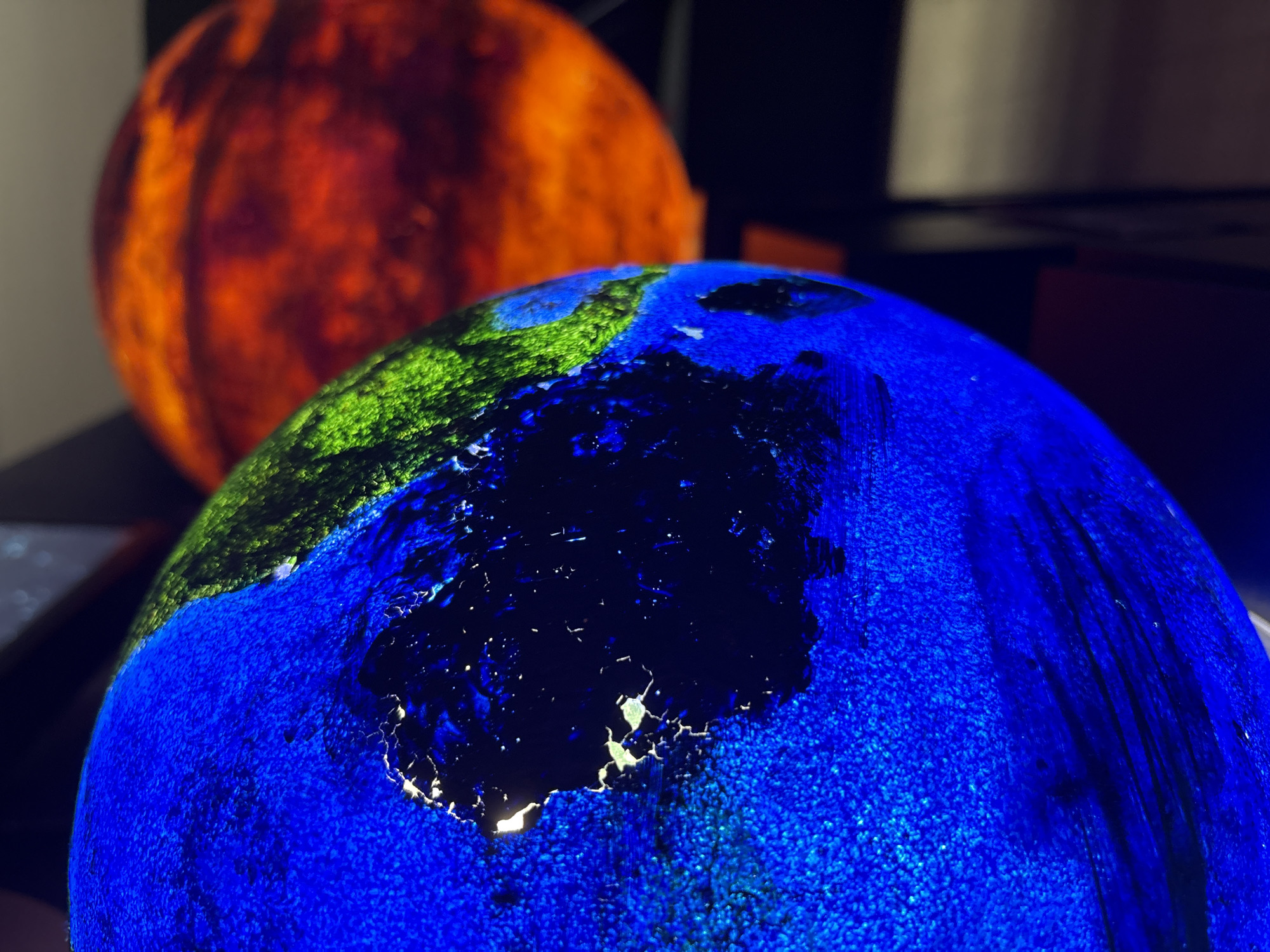 The image shows two globes. The foreground globe is blue with green continents and a large dark area that may represent a light-polluted area or a body of water at night. The background globe is orange and likely represents Mars. The globes appear to be illuminated from within. The location in Kazan, Russia is not discernible from the image.