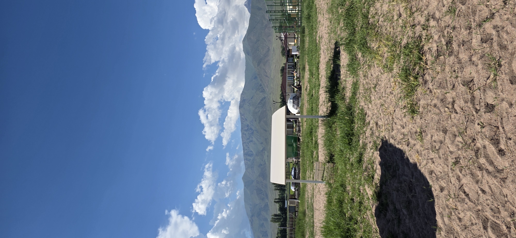The image depicts an outdoor scene with a white canopy structure in the foreground, situated on sandy ground with sparse green grass. In the middle ground, there is a grassy field with various structures including buildings with tiled roofs, a geodesic dome, and a children's playground with green netting. Beyond the field, a line of trees is visible, and behind them, a range of large, green mountains under a bright blue sky with scattered white clouds. The location is identified as Choktal, Kyrgyzstan. No people are present in the image.