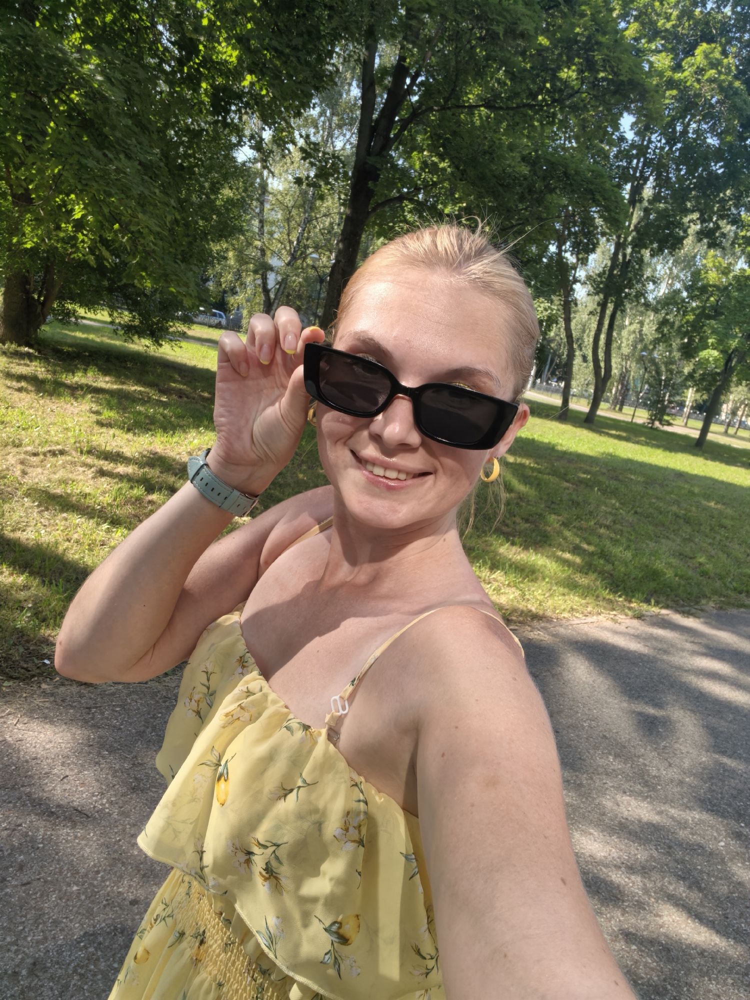 In a sunlit outdoor setting, a fair-skinned woman with blonde hair pulled back is captured in a selfie. She is wearing large black sunglasses and a yellow sundress with a floral and lemon print, adorned with ruffles. A light blue watch is visible on her left wrist. Her right hand is raised, adjusting the sunglasses. The background features a park with lush green trees and grass, suggesting a warm day. The woman is smiling, and her overall demeanor appears relaxed and happy. The scene is consistent with a park location in Sterlitamak, Russia, during a pleasant season.