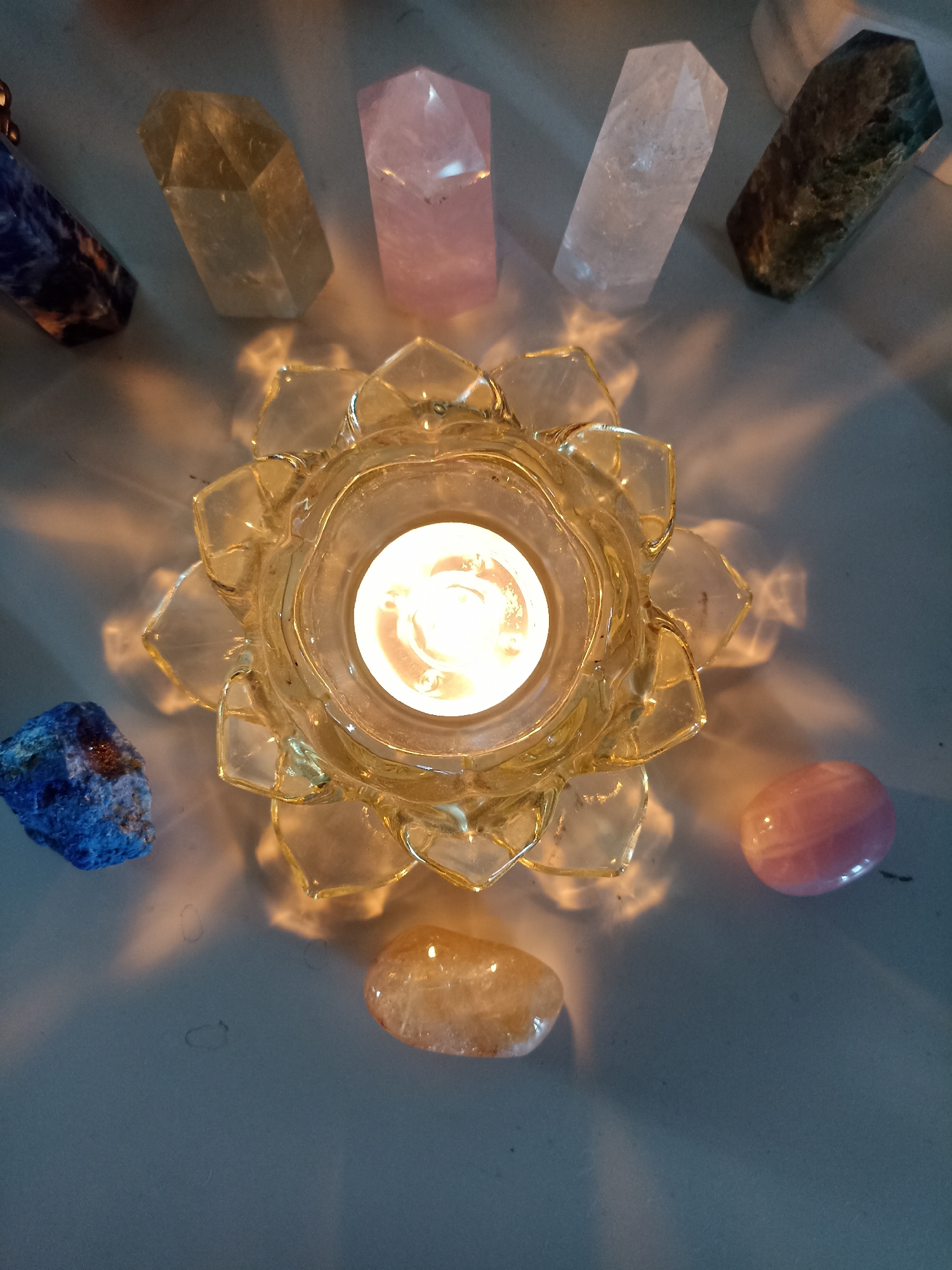 The image shows a lit candle in a lotus-shaped glass holder, surrounded by several polished crystals. The crystals appear to be of various types and colors, including blue, yellow, pink, and clear. The scene is illuminated by the candle, casting shadows and highlights on the surface. The background is a neutral-colored surface, possibly a table or shelf. There are no people visible in the image. The image does not contain any information that would allow for the determination of its location in the city of Novosibirsk, Russia.