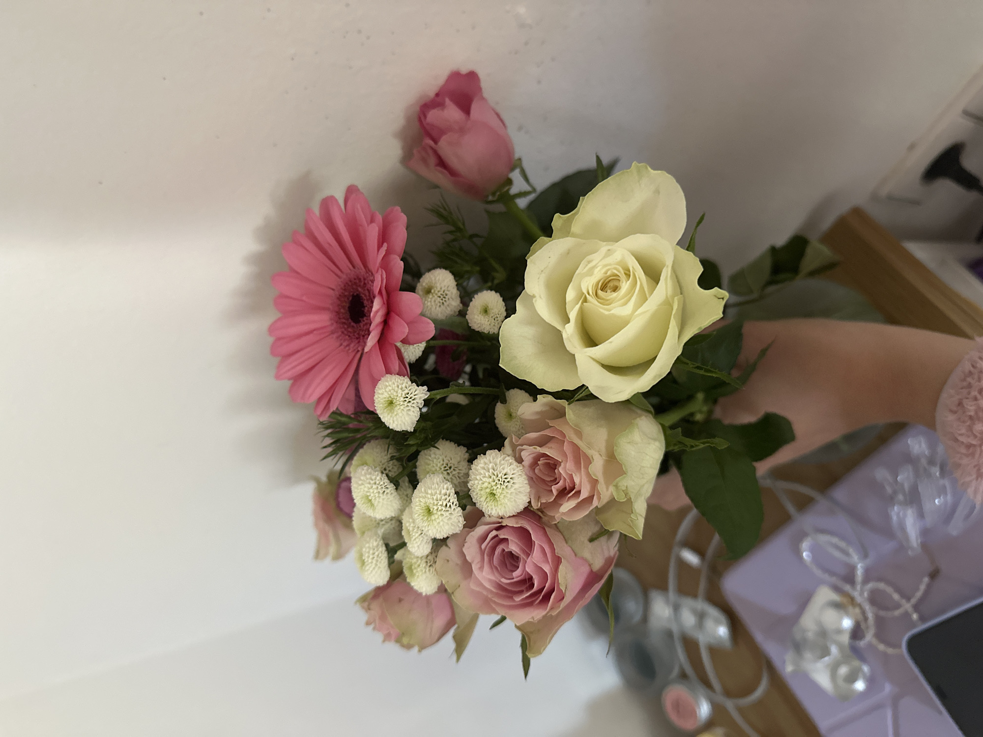 A close-up image shows a small bouquet of flowers being held. The flowers include a pink gerbera daisy, a white rose, pink roses, and small white pom-pom-like flowers. The background is a plain white surface. Other objects such as earrings, jewelry, a cell phone, and a light purple tray are in the blurry background, suggesting it may be an indoor space such as a bedroom. Based on the items and colors, this may be a personal space for a younger female. It is not possible to confirm the city or country of the image.