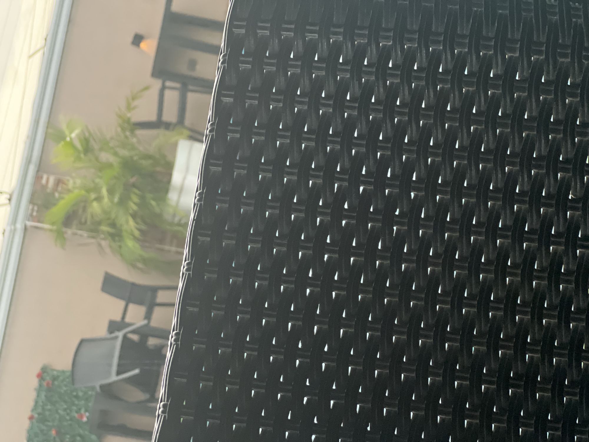 The image presents an outdoor scene, with a significant portion of the frame filled by a close-up of a dark, intricately woven material, likely rattan or similar synthetic wicker, which appears to be part of a chair or screen. This material occupies the right side of the image, showcasing a repetitive basket-weave pattern with small, bright gaps allowing light to pass through.
In the blurred background, an outdoor patio or garden area unfolds. A light-toned wall or building exterior provides the backdrop, against which various elements are visible. Lush green foliage, resembling a plant or small tree, stands in the mid-ground. Several pieces of dark outdoor furniture, including chairs, are arranged, with some appearing stacked or leaning. Further back, what seems to be a table surrounded by chairs and an overhead structure with a light fixture can be discerned. In the lower-left corner, a patch of green, possibly a hedge or artificial turf, is accented with small red details, potentially flowers.
The scene is well-lit, indicating it is daytime, likely morning or afternoon, with soft, diffused light and an absence of harsh shadows. There is no discernible human activity. The overall setting suggests a calm and quiet outdoor leisure space, such as a restaurant patio, hotel terrace, or a private garden in a warm climate like Abuja, Nigeria. No visible text is present in the image.
