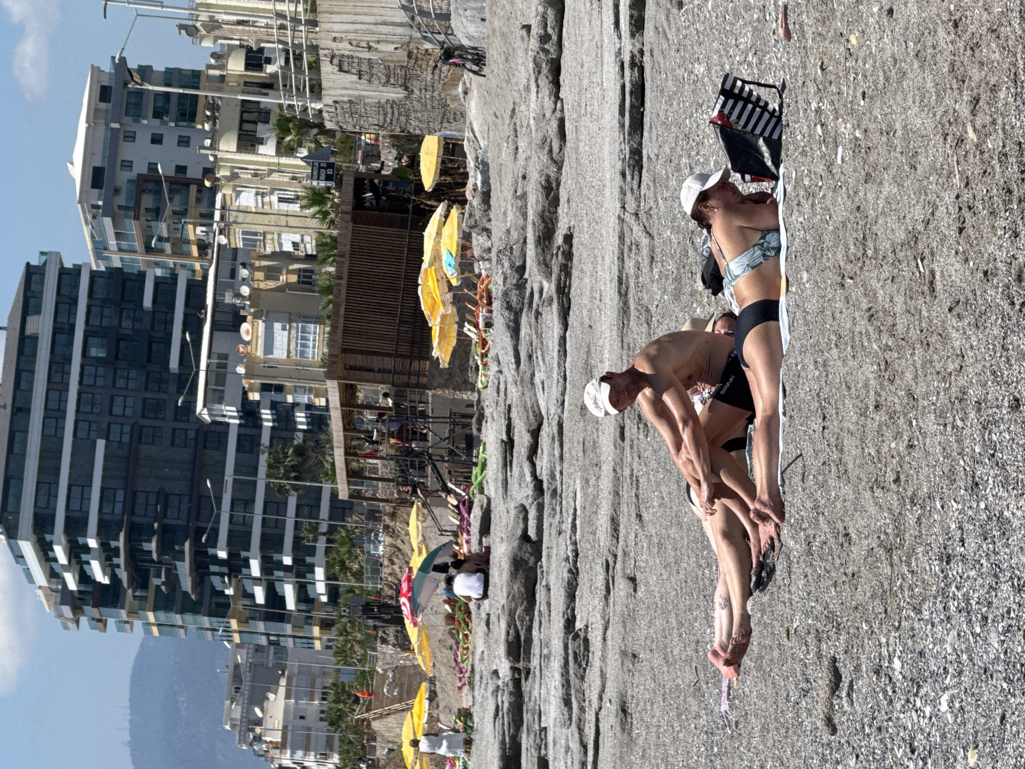 A beach in an unknown city features beachgoers sunbathing on a rocky beach. Behind the beach and elevated above it are restaurants and shops. Behind that are high-rise buildings.