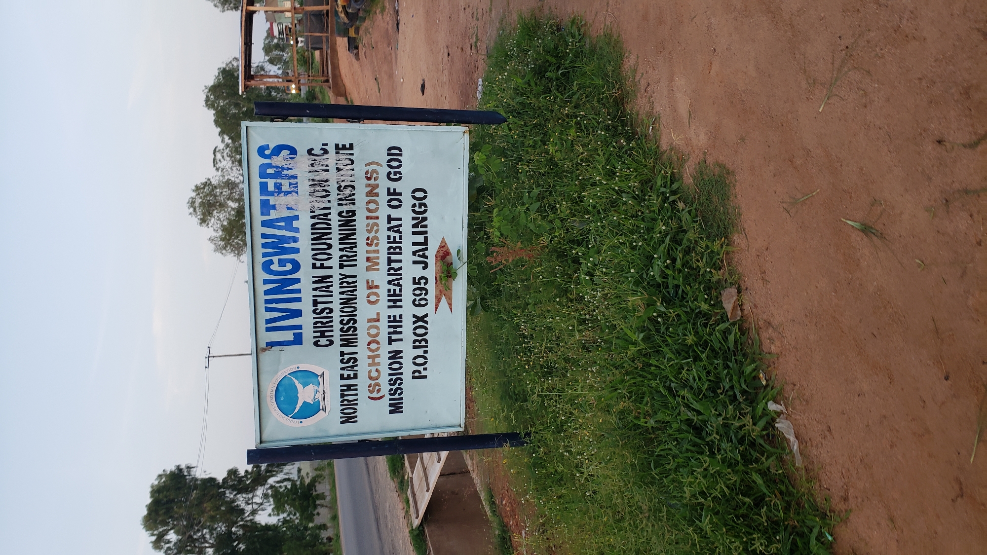 A light blue sign with dark blue and black lettering stands on a dirt path beside a grassy area. The sign reads "LIVINGWATERS CHRISTIAN FOUNDATION INC. NORTH EAST MISSIONARY TRAINING INSTITUTE (SCHOOL OF MISSIONS) MISSION THE HEARTBEAT OF GOD P.O.BOX 695 JALINGO". A logo of a dove over a globe is to the left of the text. A small red and yellow arrow graphic is below the address. The sky is overcast and the area appears to be rural or semi-rural.