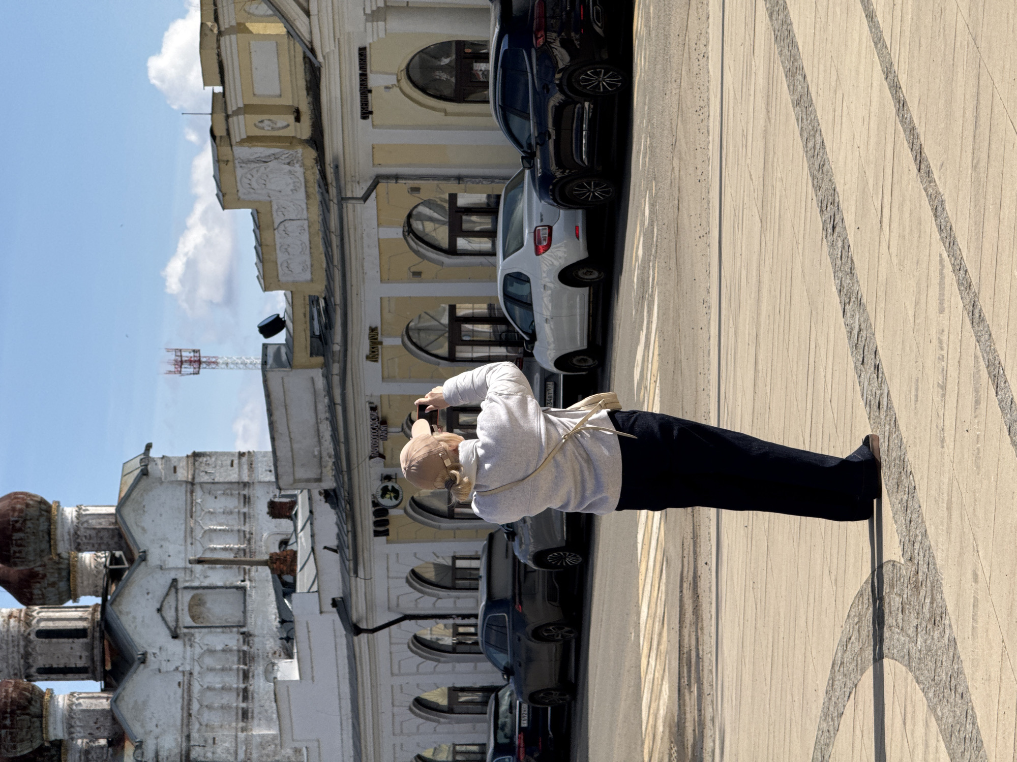 A woman stands on a paved surface taking a photograph. She wears a light gray sweatshirt, dark pants, brown shoes, and a light tan cap. She is holding a smartphone up, presumably to take a picture of something in front of her.
Behind the woman are several parked cars and a building with multiple arched windows. In the background, to the left, is an older-style building with multiple domed roofs.
The photograph was taken in Novo-Nikol'skoe, Russia.