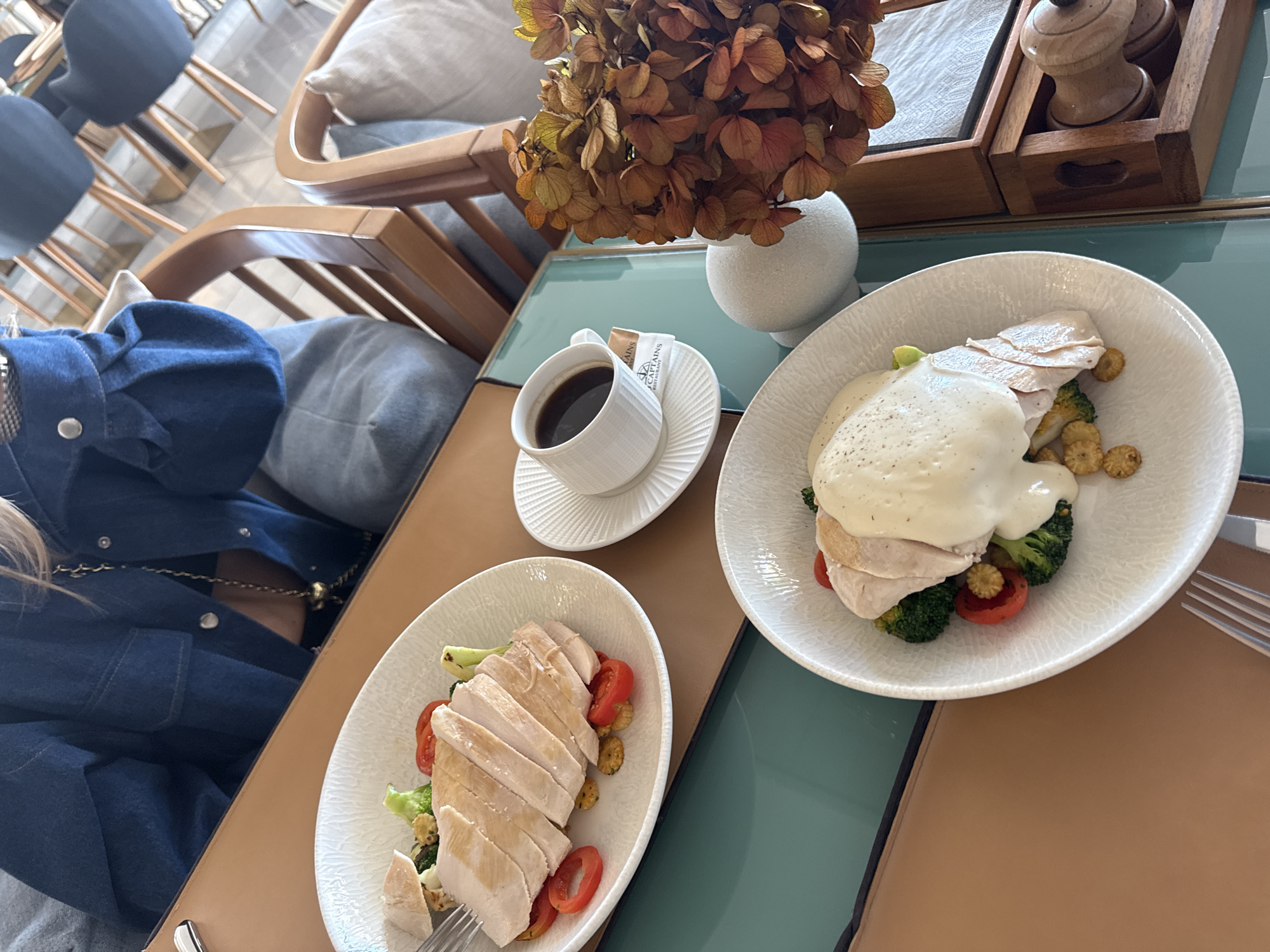The scene presents a meal at an establishment in Unknown, Unknown. A woman in a denim jacket is seated at a table. The table is set with two white plates containing sliced chicken breast accompanied by vegetables, along with a cup of coffee on a saucer. A vase with dried hydrangeas sits centrally on the table, next to a wooden tray holding condiments. Utensils are laid out for the meal. The surrounding environment includes chairs and a lounge, indicating a comfortable, casual setting.
