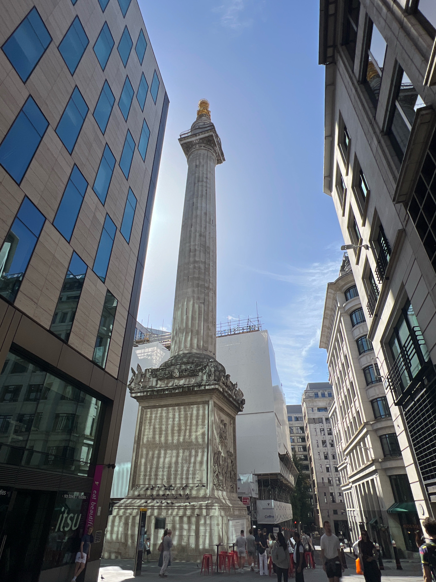 The image shows the Monument in London, United Kingdom, a tall Doric column erected in 1677 to commemorate the Great Fire of London. The Monument is situated in the financial district, with modern buildings surrounding it.
In the foreground, to the left, is a contemporary building with a facade of beige stone and diamond-shaped blue glass windows. The building has a reflective surface, showing the sky and surrounding architecture. The lower part of this building features signage for "itsu" and "eat beautiful," indicating a restaurant.
To the right of the Monument, a more traditional stone building with multiple floors and dark windows rises. Further down the street, other buildings of varying architectural styles are visible, including some under scaffolding, suggesting ongoing construction or renovation.
The Monument itself is a prominent feature, with its tall, fluted column topped by a gilded copper urn. The base of the monument is adorned with intricate carvings and inscriptions. The sun is bright, casting shadows and creating a lens flare effect in the upper portion of the image.
Several people are walking in the street around the Monument and on the sidewalks. They are dressed in casual summer clothing. Some are walking in groups, while others are individuals. Red stools are visible near the "itsu" restaurant.
The sky is blue with scattered white clouds. The overall scene depicts a busy urban environment in London, blending historical landmarks with modern development.