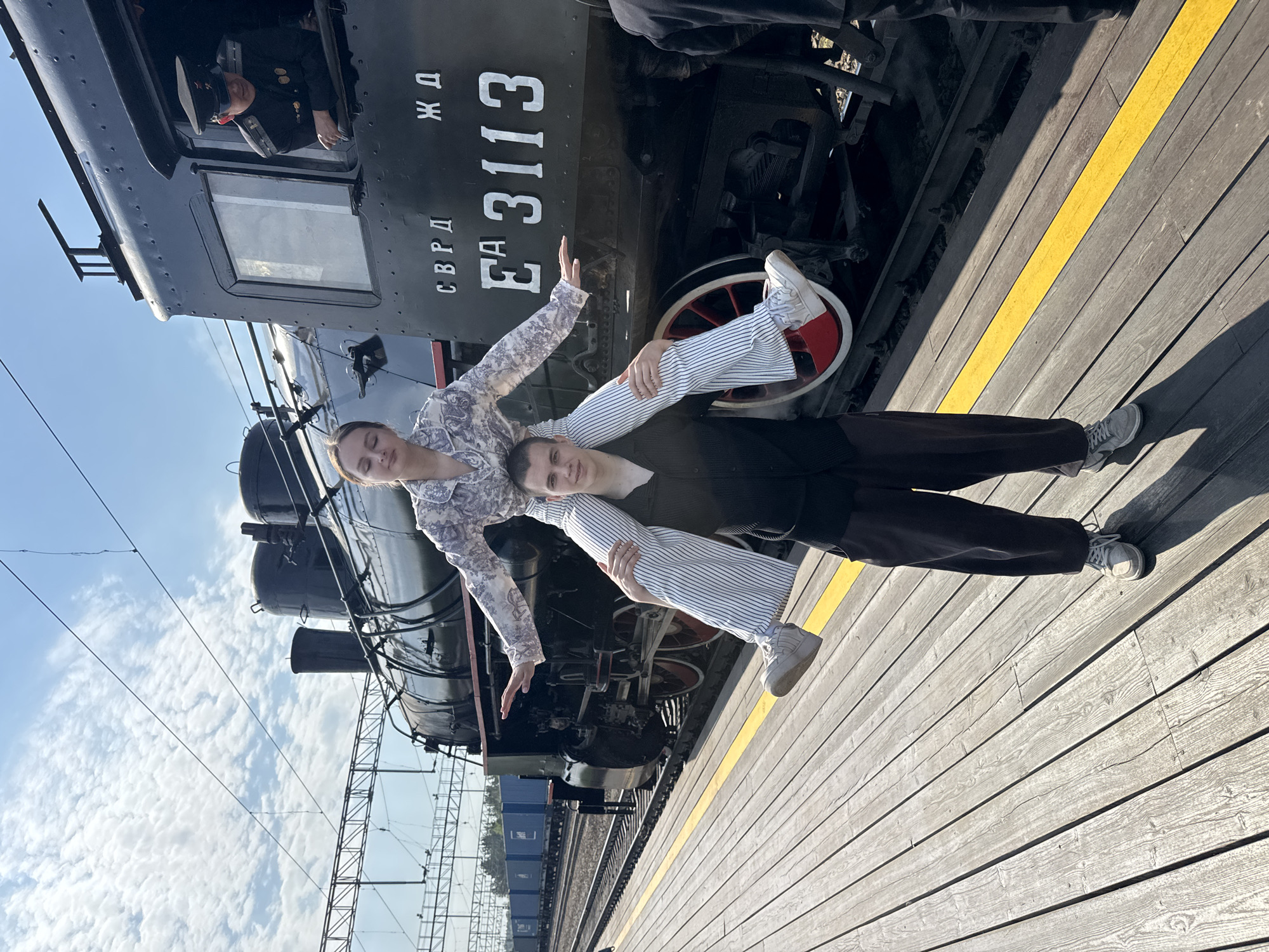 A man supports a woman horizontally over his shoulders in front of a black steam locomotive at the Yekaterinburg railway station in Russia. The locomotive has the Cyrillic letters "СВРД" and "ЖД" painted on it, along with the number "ЕА 3113". A man in a conductor's uniform peers out from the train's cab. The couple is on a wooden platform with a yellow safety line. A train is visible on the tracks in the background. The sky is light blue with scattered clouds.