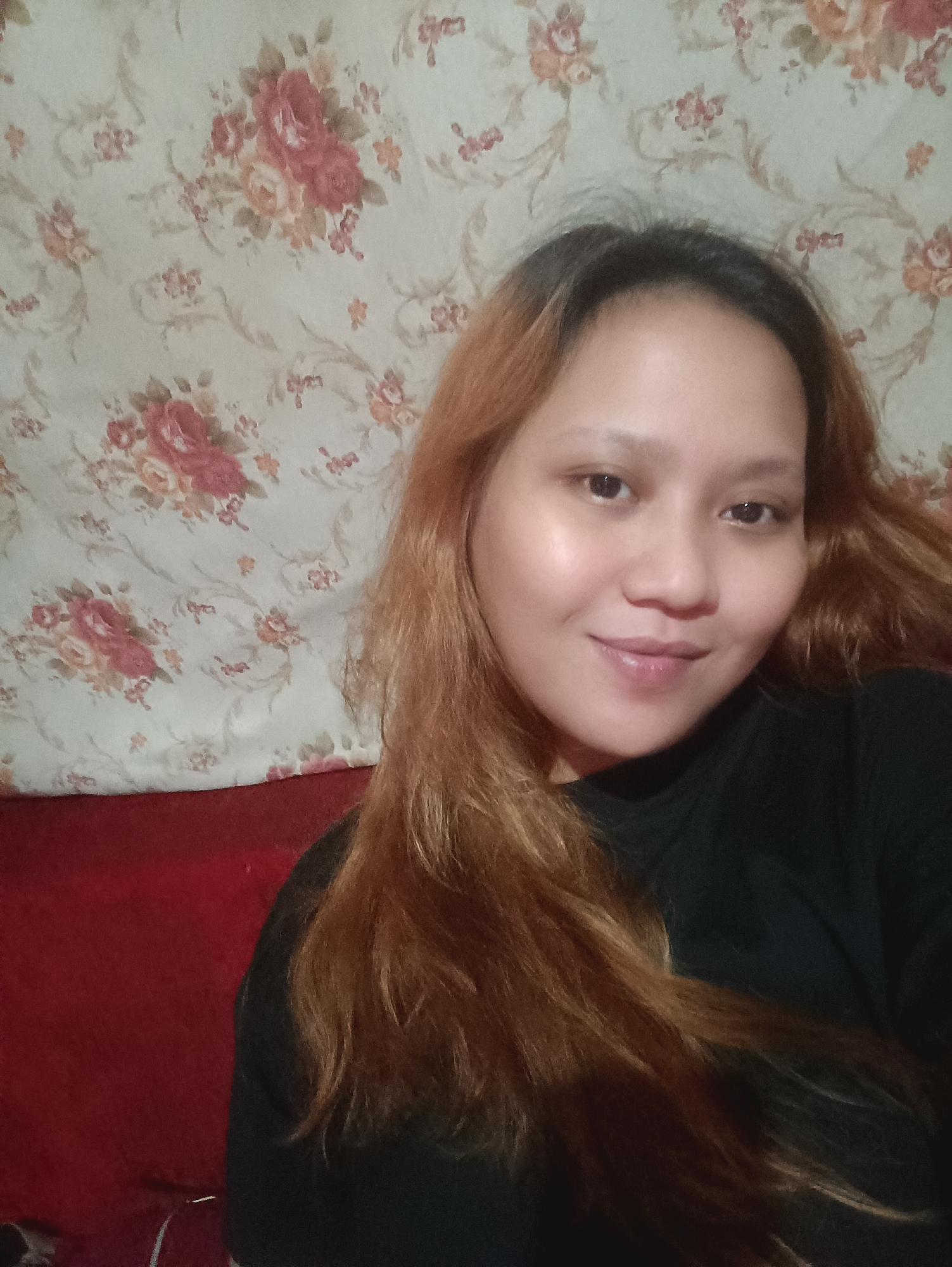 A young woman with auburn hair is taking a selfie in front of a floral patterned wall. She is wearing a black top. To her left, a red cushioned surface is visible. The scene appears to be indoors.