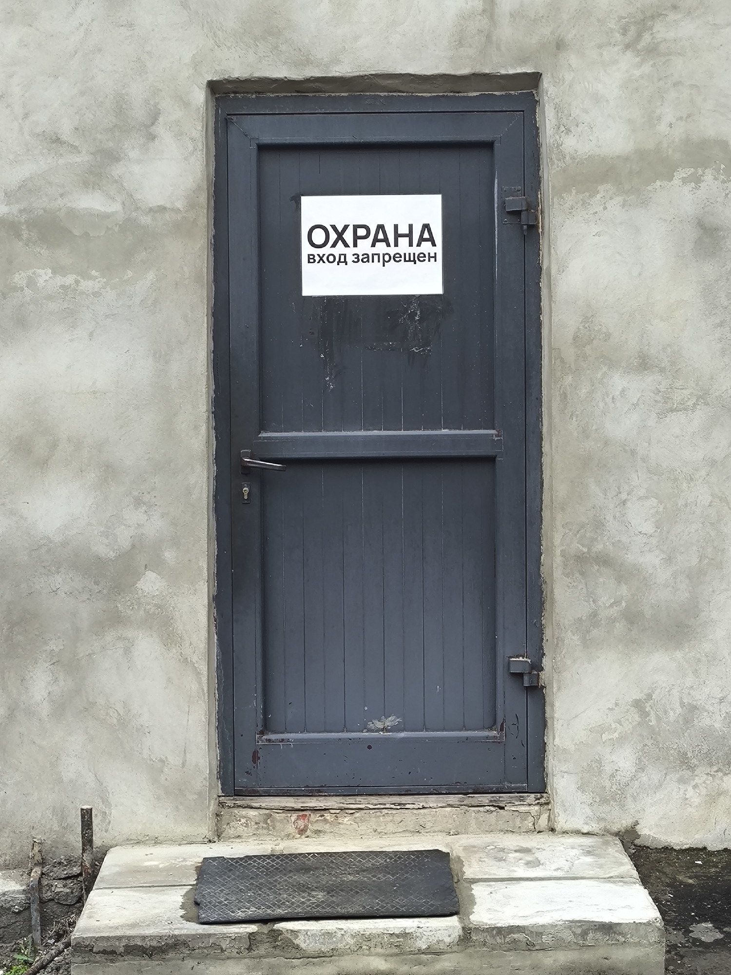 A gray metal door with a sign in Russian, "ОХРАНА вход запрещен" (SECURITY, entry prohibited), is set into a textured gray wall. The door has a horizontal bar and a handle. A gray rubber mat sits on the concrete step in front of the door. Some vegetation is visible near the base of the wall. The location of Moscow, Russia, is not evident from the image.