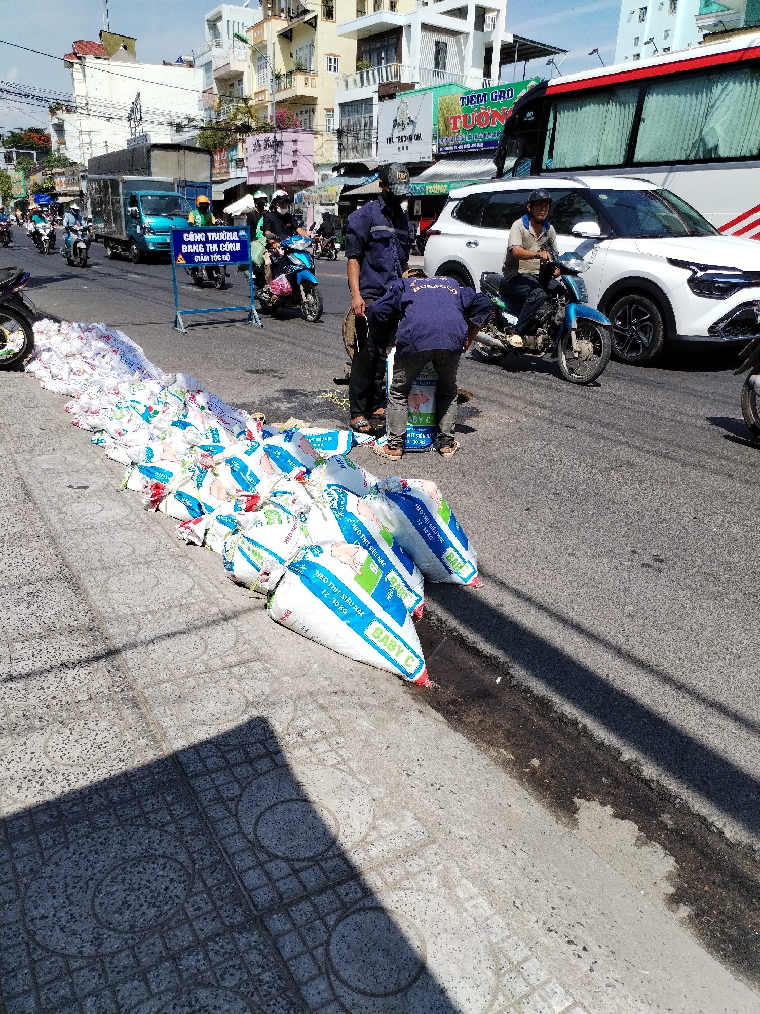 On a sunny day in Bac Nha Trang Ward, Vietnam, a street scene unfolds with a prominent line of white sacks, possibly containing animal feed, stretching along the sidewalk and partially into the roadway. The sacks are emblazoned with blue and white lettering, including "BABY C" and "HEO THIT SIEU NAC" (super lean pork), with some indicating a weight range of 12-30 KG. A few of the sacks are open, revealing white contents.
Several individuals are present. Two men in blue work shirts and grey pants are near the sacks. One man, facing away from the camera, appears to be interacting with the sacks, while another man in a dark blue shirt and dark pants stands closer to the road. A third man, wearing a light-colored polo shirt and dark pants, is seated on a blue motorcycle, observing the scene. A fourth person, dressed in a black shirt and black pants, is also on a motorcycle, positioned behind the man in the polo shirt. Two other individuals on motorcycles, one in a green shirt and helmet and another in a yellow helmet, are further down the street. A teal truck with a covered bed is parked on the road, and a white SUV is also present. In the background, a large white bus with red trim is visible, along with a white building displaying signage and other structures. A blue and white directional sign with Vietnamese text and a traffic symbol is placed on the sidewalk. The environment includes a paved road with a drainage ditch, a tiled sidewalk, and overhead power lines.