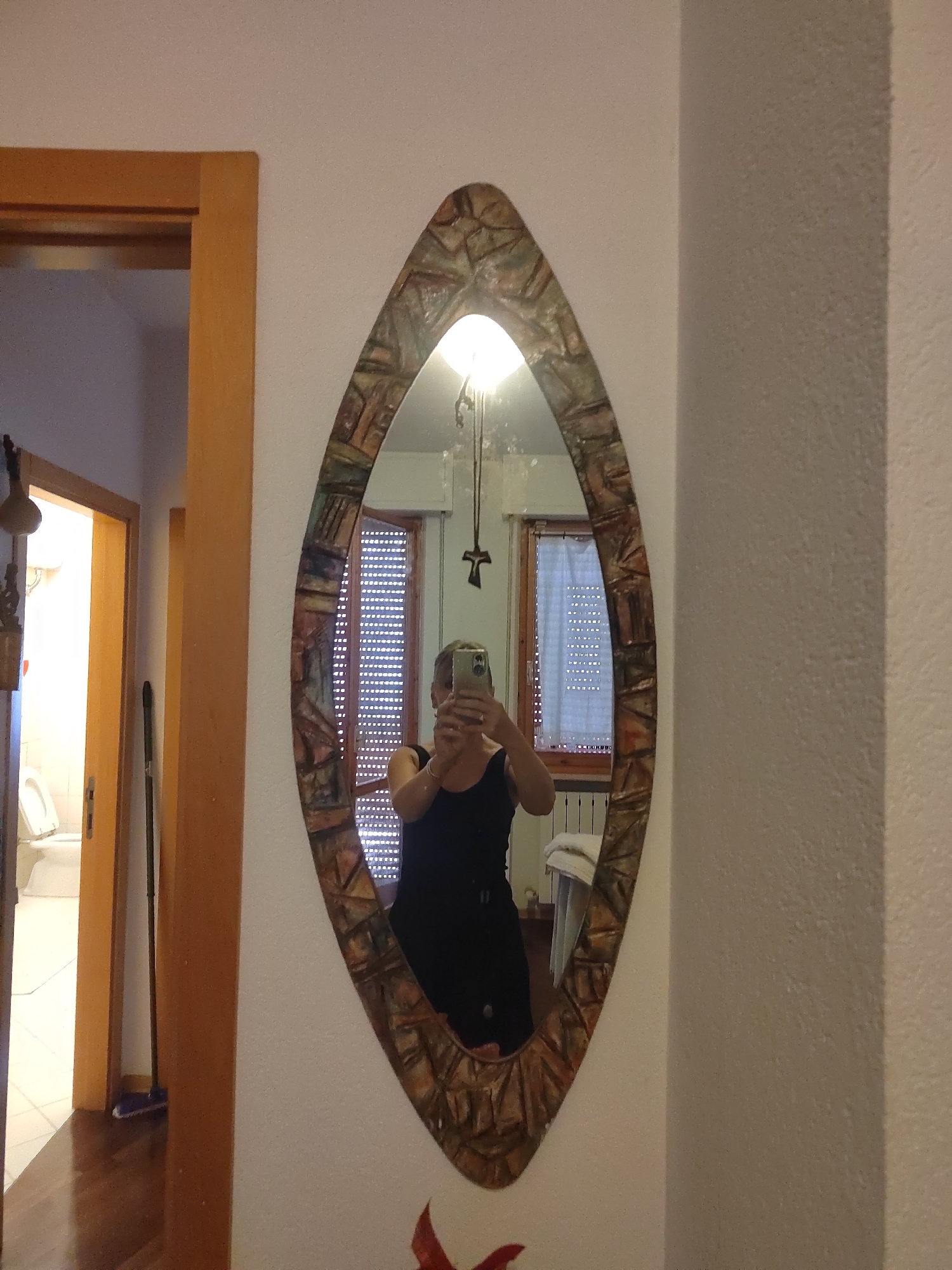 The image depicts an interior scene with a person taking a selfie in an oval-shaped mirror. The mirror has a decorative frame with a mosaic-like pattern of textured pieces in earthy tones. The person is wearing a black sleeveless top and holding a silver smartphone. In the reflection, a window with horizontal blinds is visible, and a white towel is draped over a railing in the background. To the left of the mirror, a doorway leads to another room, revealing a toilet and a door ajar. A mop with a blue head stands near the doorway. The walls appear to be textured and painted a light color. There is a wooden doorframe on the left side of the image. The scene appears to be located in a residential setting.