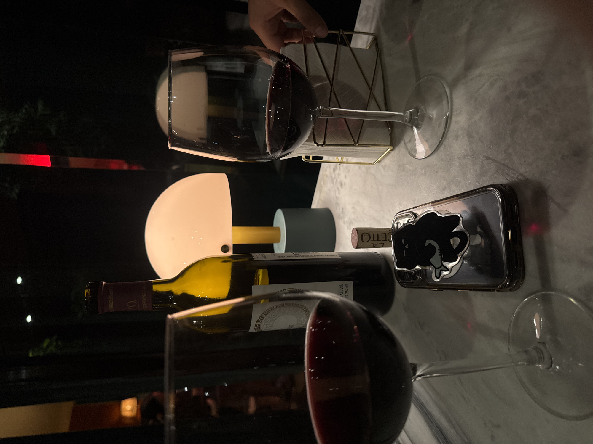 A table in Puebla, Mexico, presents a tableau of leisure. Two partially-filled wine glasses with long stems reflect light, one near the bottom of the frame and the other toward the upper-left, holding a red liquid. A wine bottle, possibly emptied, rests horizontally between the glasses, its label partially visible, along with a cork bearing the word "ETTO". A modern smartphone in a clear case, adorned with a decorative, raised cartoon silhouette, sits to the right of the bottle. A gold wire napkin holder contains white napkins, and a small lamp with a spherical white shade adds a warm glow. The table surface is a marbled white and gray. An ambiguous dark space occupies the left background, with hints of a red vertical line near the top, suggesting an interior setting. An arm and hand holding the napkin holder is visible.