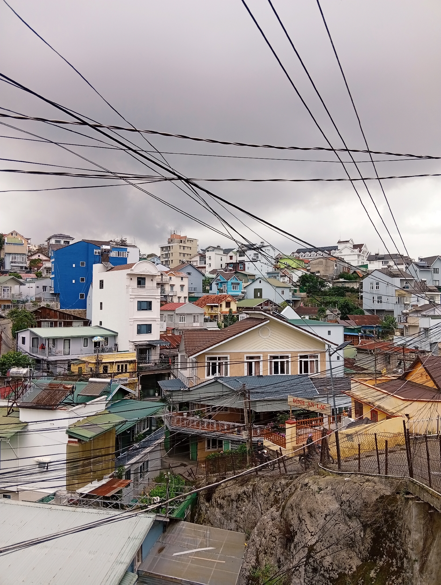 This image depicts a dense urban landscape of residential buildings in Da Lat, Vietnam. The architecture is varied, with a mix of brightly colored homes and more muted structures, many featuring pitched roofs. The foreground is dominated by a tangled network of overhead electrical wires, a common sight in Vietnamese cities, which crisscross the sky and obscure the view of the buildings behind. The buildings themselves are built closely together, with many appearing to be multi-story dwellings. Some of the houses have balconies and windows that suggest human habitation. A large, grey rock formation is visible in the lower right quadrant, partially covered by moss and vegetation, with a wire fence running along its edge. There are no discernible people actively engaged in the scene, though the presence of homes implies residents. The overall impression is one of a lively, densely populated neighborhood in a hilly terrain, with the ubiquitous electrical infrastructure adding a characteristic element to the visual composition.