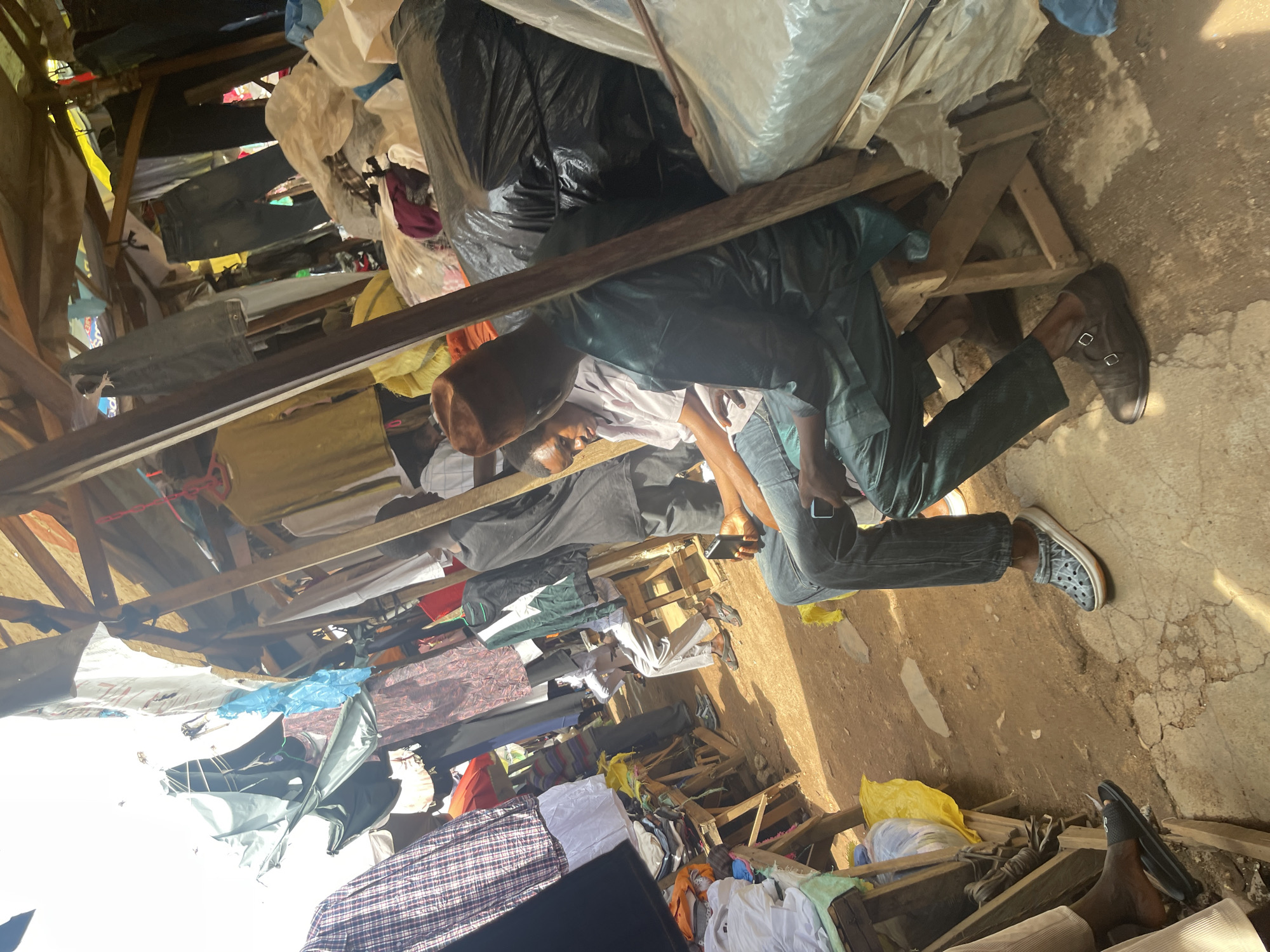 This image depicts a street market scene in Jos, Nigeria. The market appears to be outdoors, with stalls covered by tarps and wooden structures. Various clothing items are displayed, some hanging from makeshift racks and others piled on wooden tables. The ground is dusty and uneven, with concrete patches visible.
In the foreground, two individuals are seated on wooden stools. The person on the right is wearing a green traditional outfit and a brown cap, holding a mobile phone. The person on the left is wearing a white shirt and jeans, with a mobile phone in their hand as well. Their legs and feet are visible, showing their footwear. Behind them, other people are seen moving through the market, and more clothing displays are visible. Large black plastic bags are stacked to the right, suggesting merchandise storage. The overall atmosphere is that of a busy, informal marketplace.