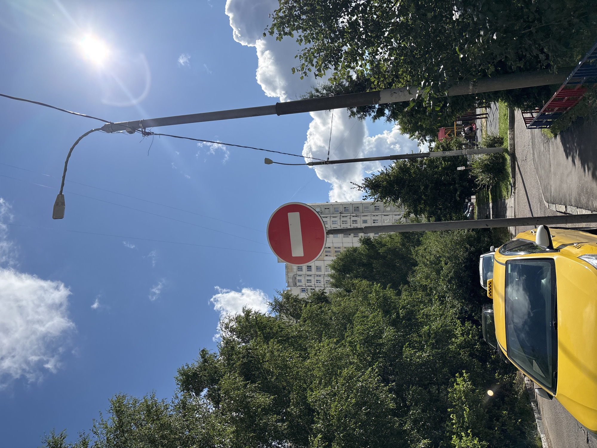The image depicts a street scene under a bright, sunny sky with scattered white clouds. A tall, light-colored apartment building is visible in the background, surrounded by lush green trees. In the foreground, a bright yellow taxi is parked on the left side of the road. A red and white traffic sign, indicating "No Entry," is prominently displayed on a pole in the center of the frame. To the right, a sidewalk with a red, white, and blue fence leads towards a children's playground. Two lampposts stand on either side of the sidewalk, with one casting a long shadow. Power lines crisscross the sky, and the sun glares brightly, creating lens flare. The overall environment appears to be a residential area in a city.
