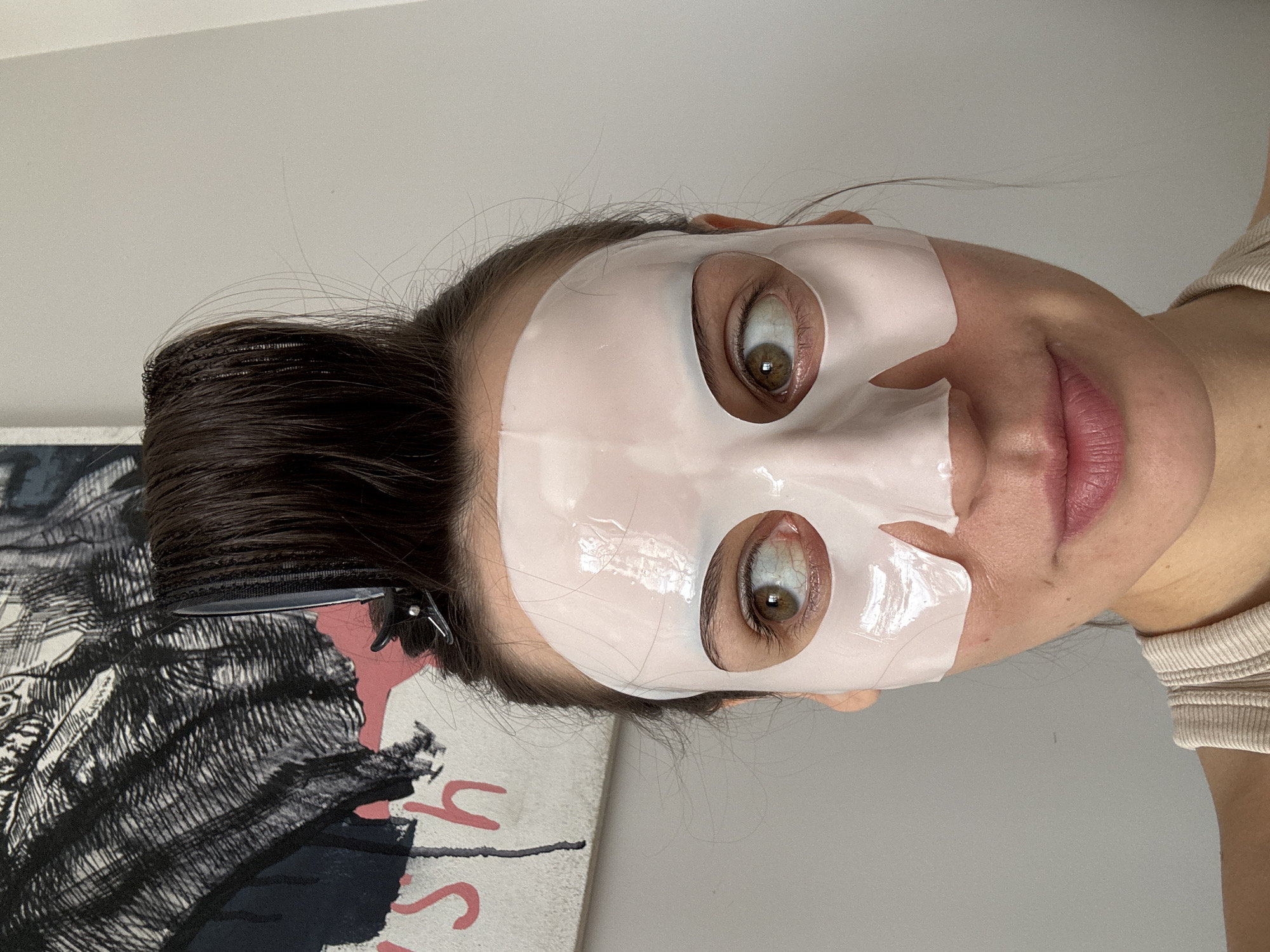 A woman with dark hair, pulled back with clips, is wearing a sheet mask on her face. The mask is white and has cutouts for her eyes. Her eyes are wide and hazel. She is wearing a ribbed top. Behind her is a painting or print with black and red elements on a white background. The background is a plain wall. There is no indication of a location in Krasnoyarsk, Russia, or any other city.