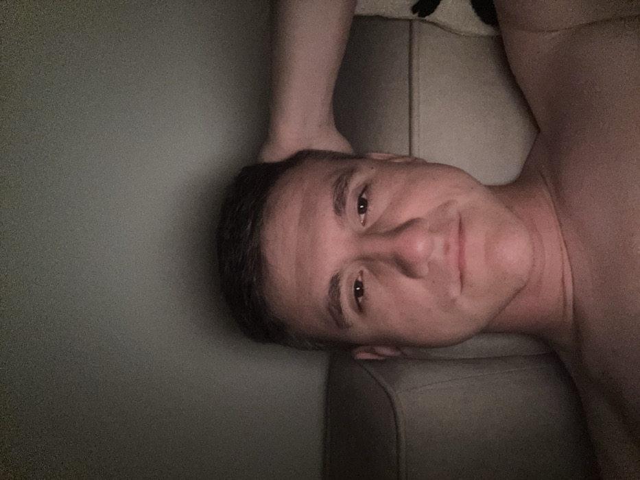 A shirtless man lies on a gray couch, his head resting on the back of the sofa. His arm is raised, the hand touching the side of his head. He gazes directly at the viewer with a slight smile. The lighting is dim, casting subtle shadows across his face and body. A section of the wall is visible on the left side.