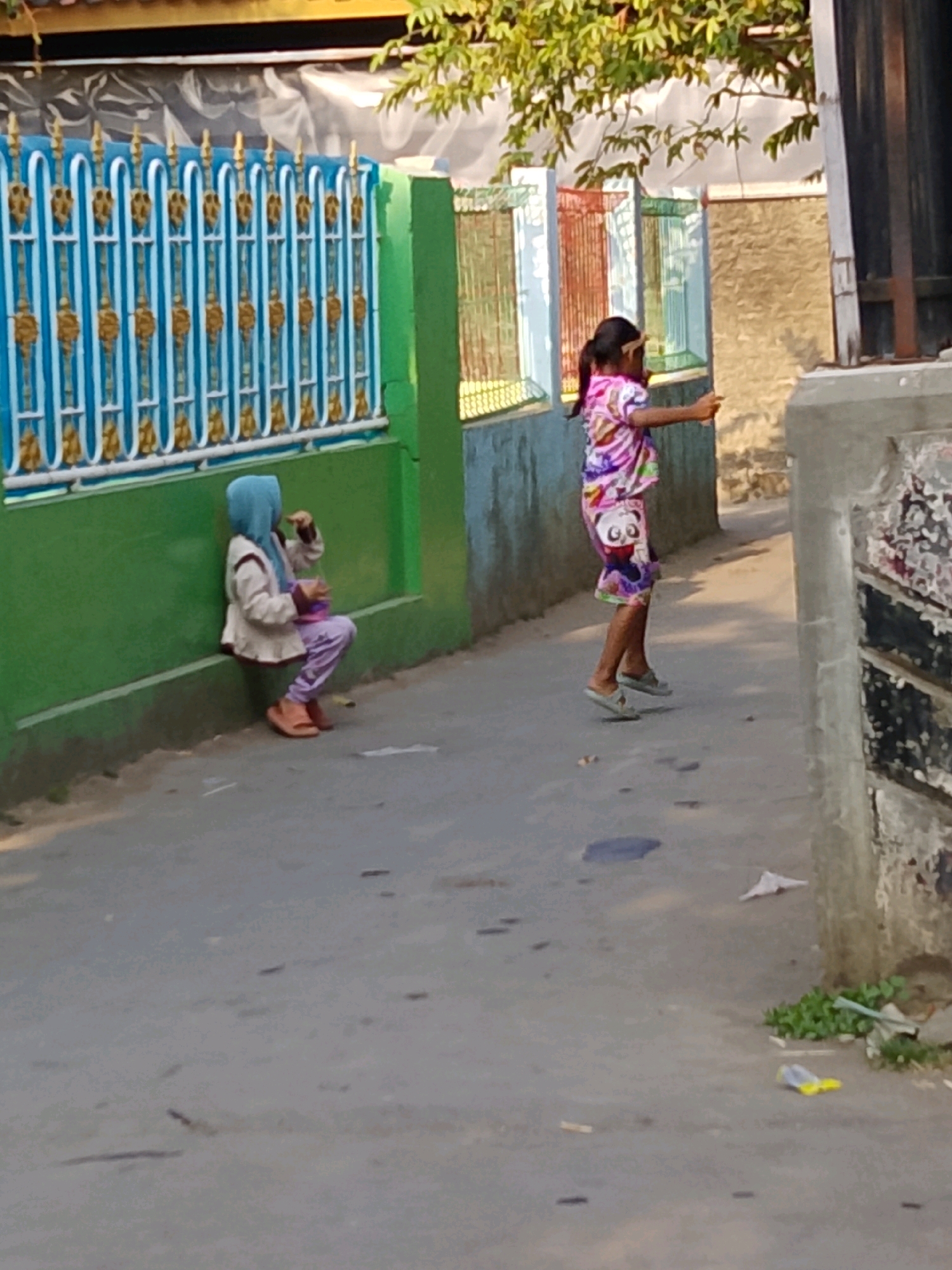 The image depicts an outdoor scene, likely in a residential alley or street, in Cirebon, Indonesia. Two young girls are the primary subjects. The girl on the left is seated on the ground, leaning against a green wall with a blue and gold ornate fence. She is wearing a light-colored jacket, a blue hijab, and purple pants, and appears to be holding a small object in her hand. The girl on the right is standing and facing away from the viewer, wearing a colorful, patterned shirt and skirt with a panda design. She has her hair pulled back in a ponytail and is reaching her right hand out, possibly holding a small item. The environment includes a paved surface, buildings with varying textures and colors, and some greenery. The lighting suggests it is daytime, possibly late afternoon, given the shadows.